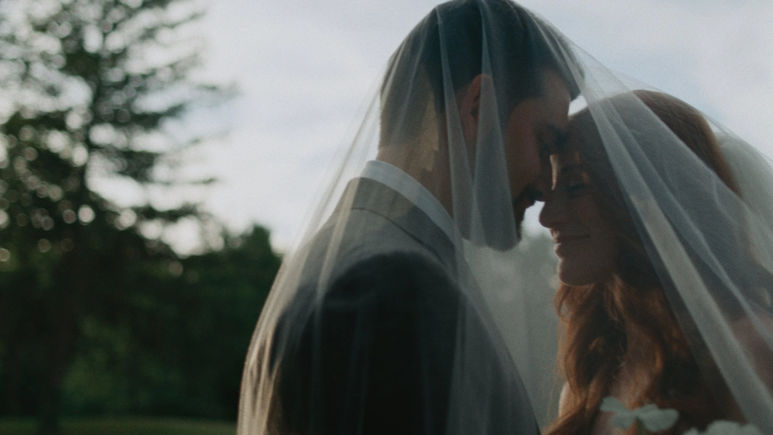 A bride and groom standing close under a veil outdoors, with trees in the background, touching foreheads and smiling.