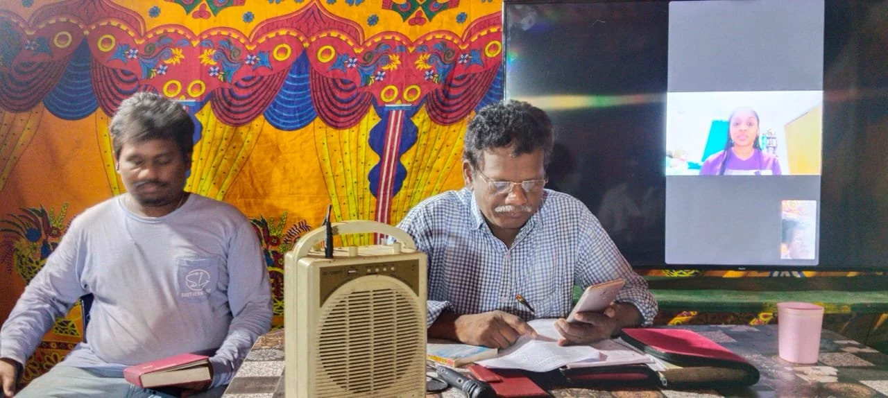 Two men sitting at a table with a colorful, patterned backdrop. One is looking at a phone, and the other has a book. A television screen nearby shows a video call with a person speaking, and a radio is placed on the table.