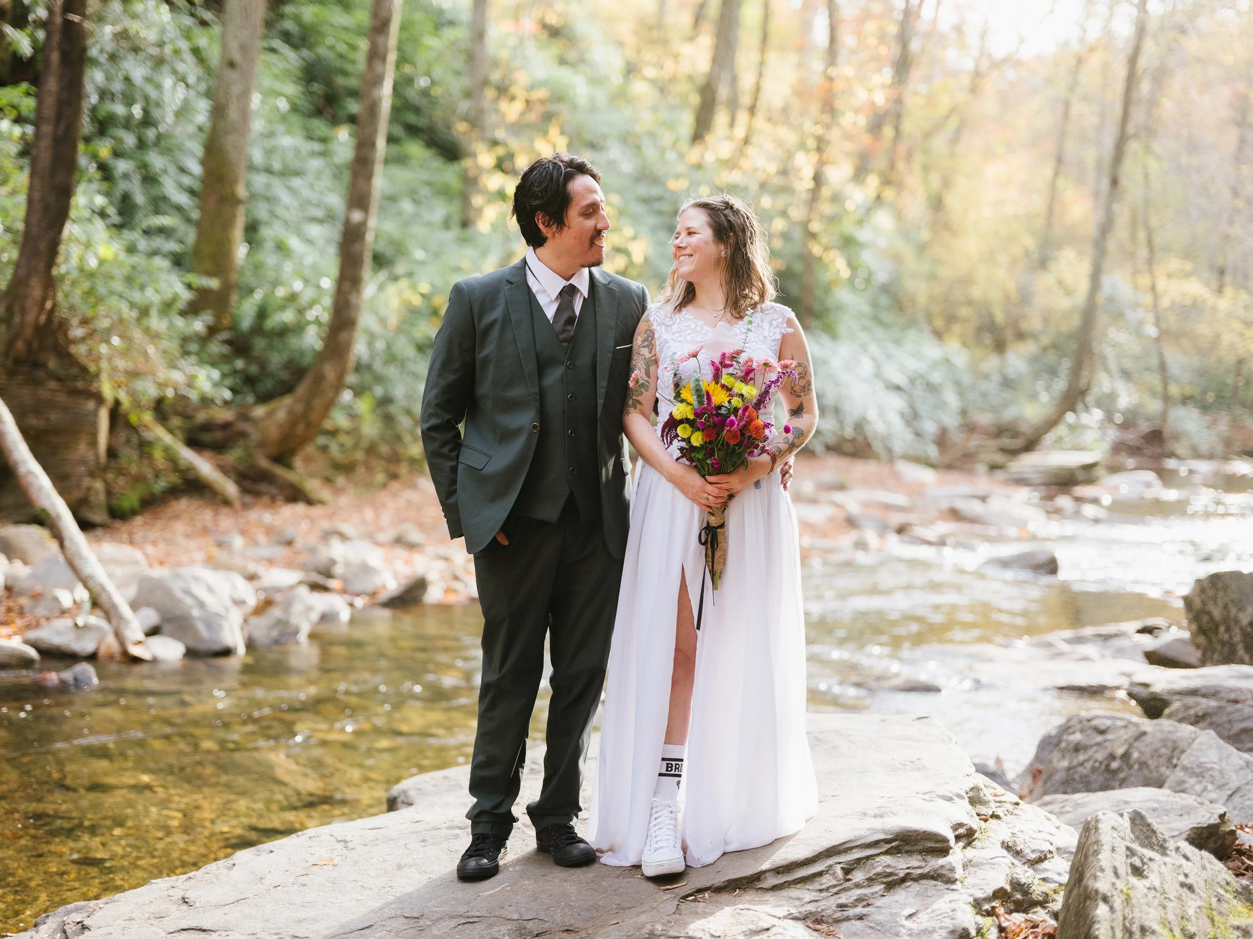 A couple dressed in wedding attire standing on a large rock beside a stream in a forest, smiling at each other. The woman is holding a colorful bouquet of flowers.