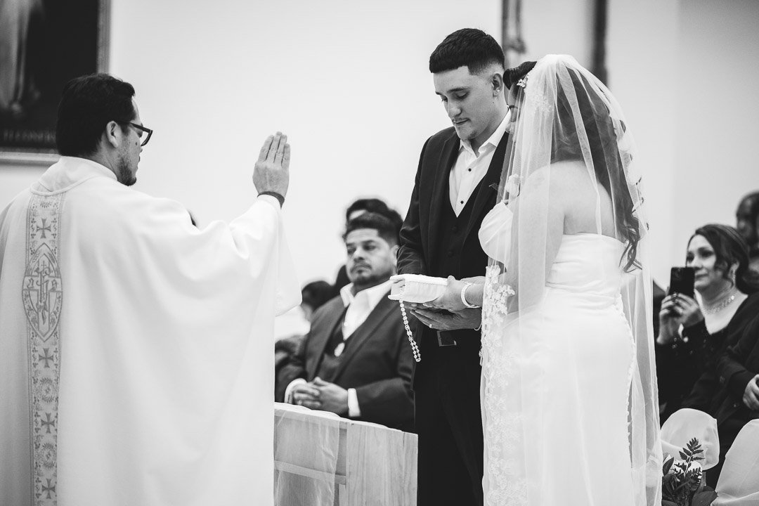 A wedding ceremony with a priest conducting vows between a groom and bride, with guests observing.