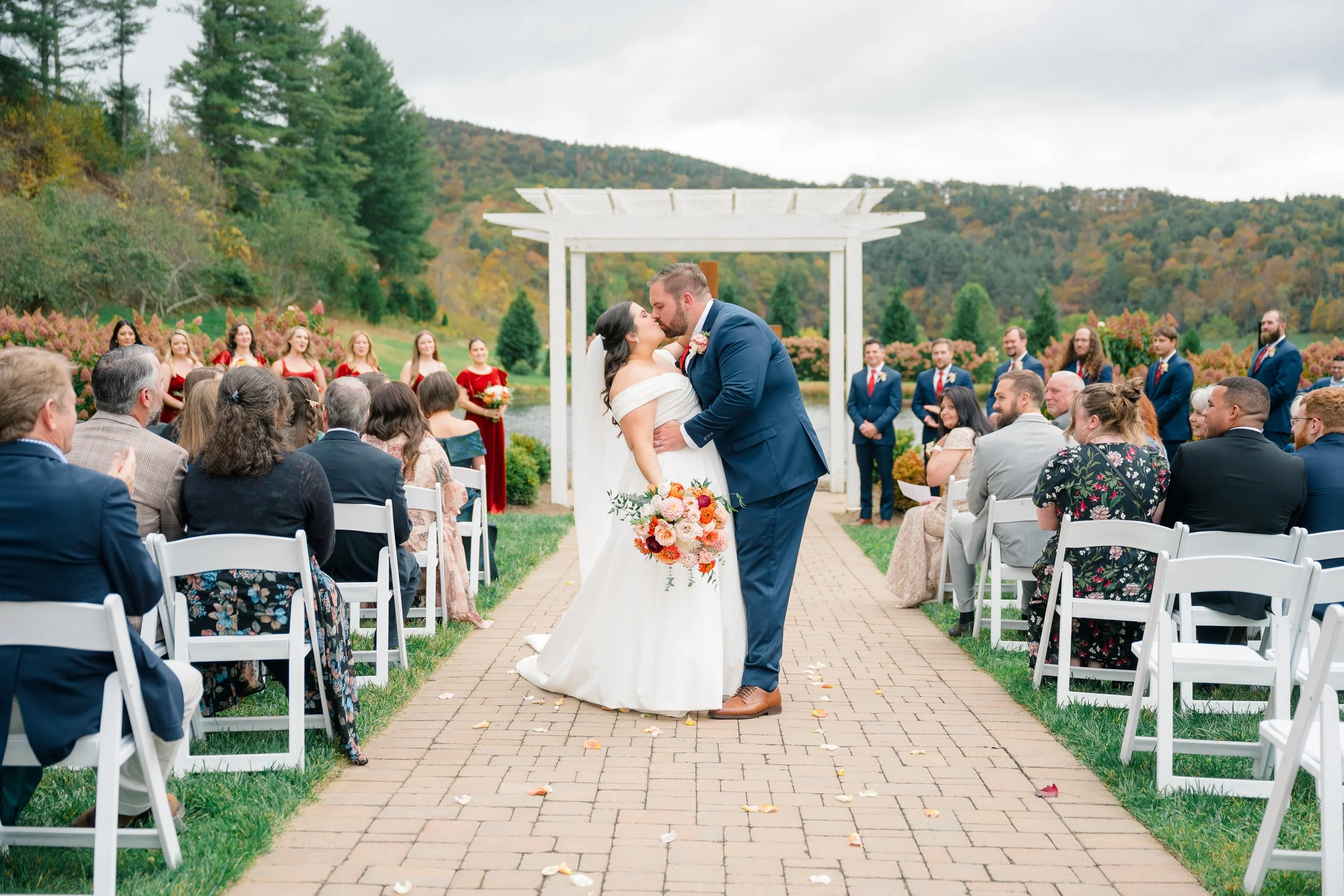 A bride and groom kissing during their outdoor wedding ceremony in a scenic natural setting with hills and trees in the background. The bride is holding a bouquet of pink, orange, and white flowers, and guests are seated on white chairs on either sid