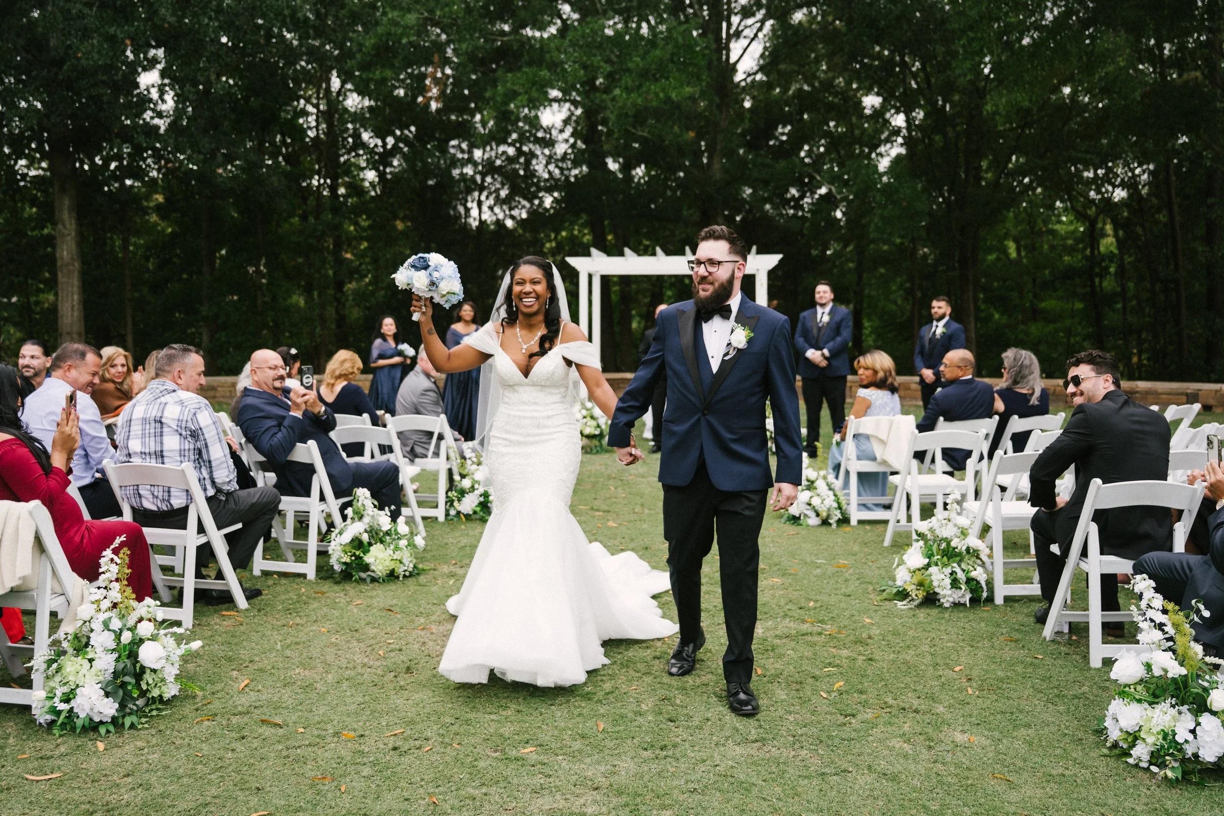 A newly married couple walks down the aisle at an outdoor wedding. The bride holds a bouquet of white and blue flowers, and the groom is dressed in a navy blue tuxedo. Guests sit on white chairs decorated with white flowers, with some taking photos. 