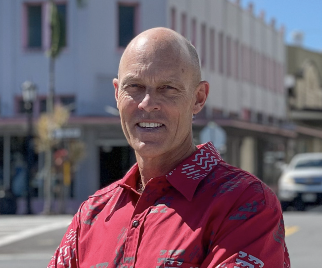 A man in a red patterned shirt standing outdoors in front of a building with pink and white trim, with a blurred background and a car on the street.