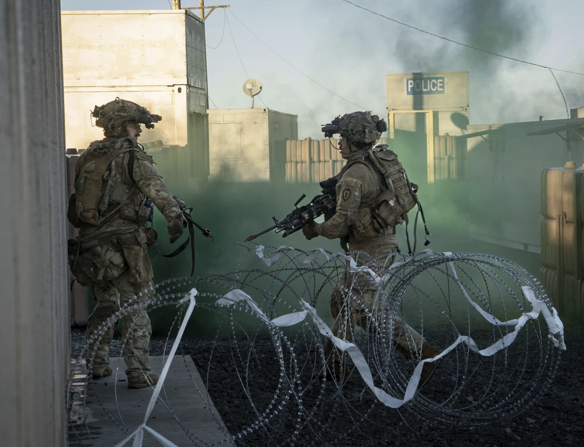 Two soldiers in combat gear standing behind concertina barbed wire in a military setting with a police sign in the background and green smoke or tear gas in the air.