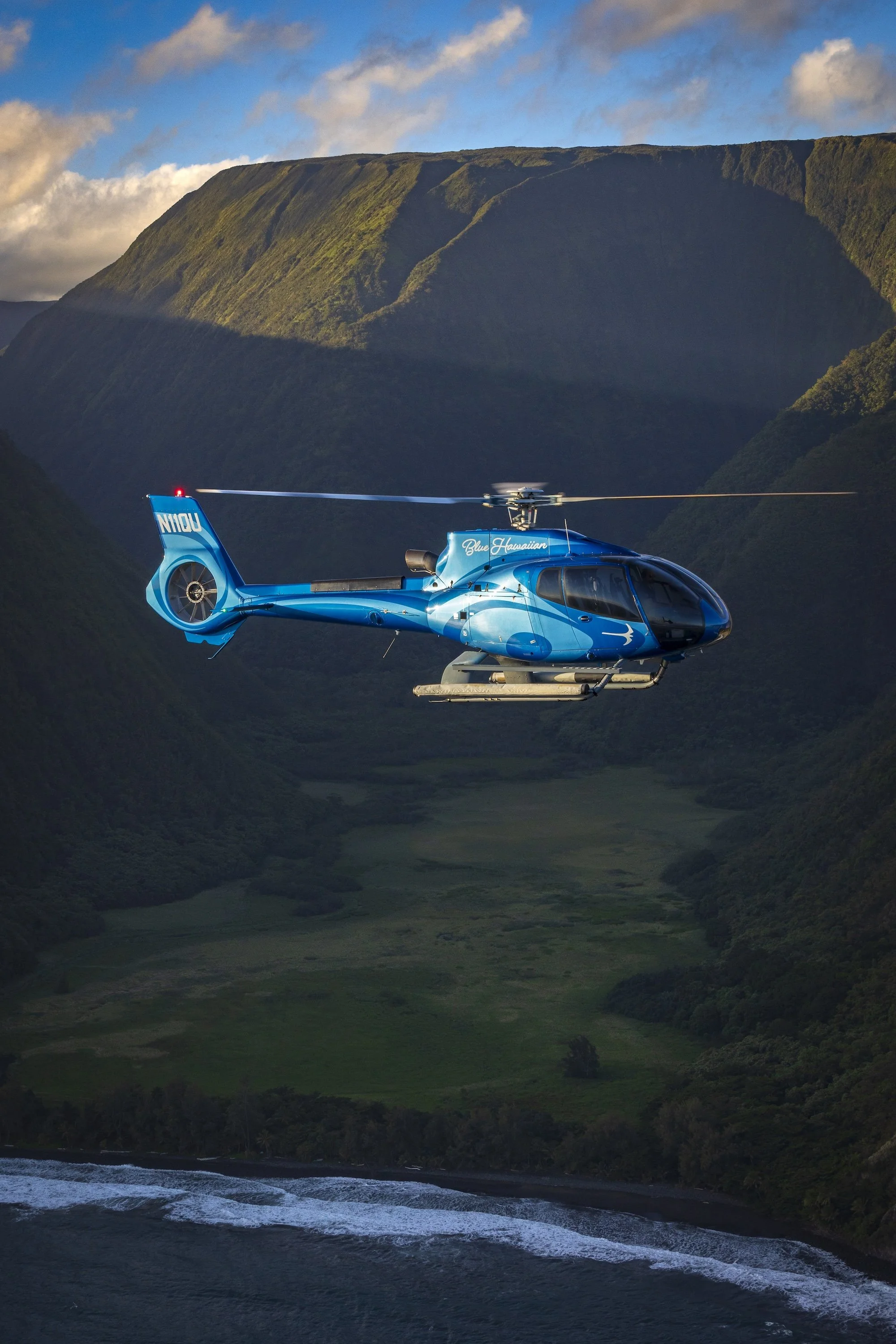 A blue helicopter flying over a lush green valley surrounded by mountains.