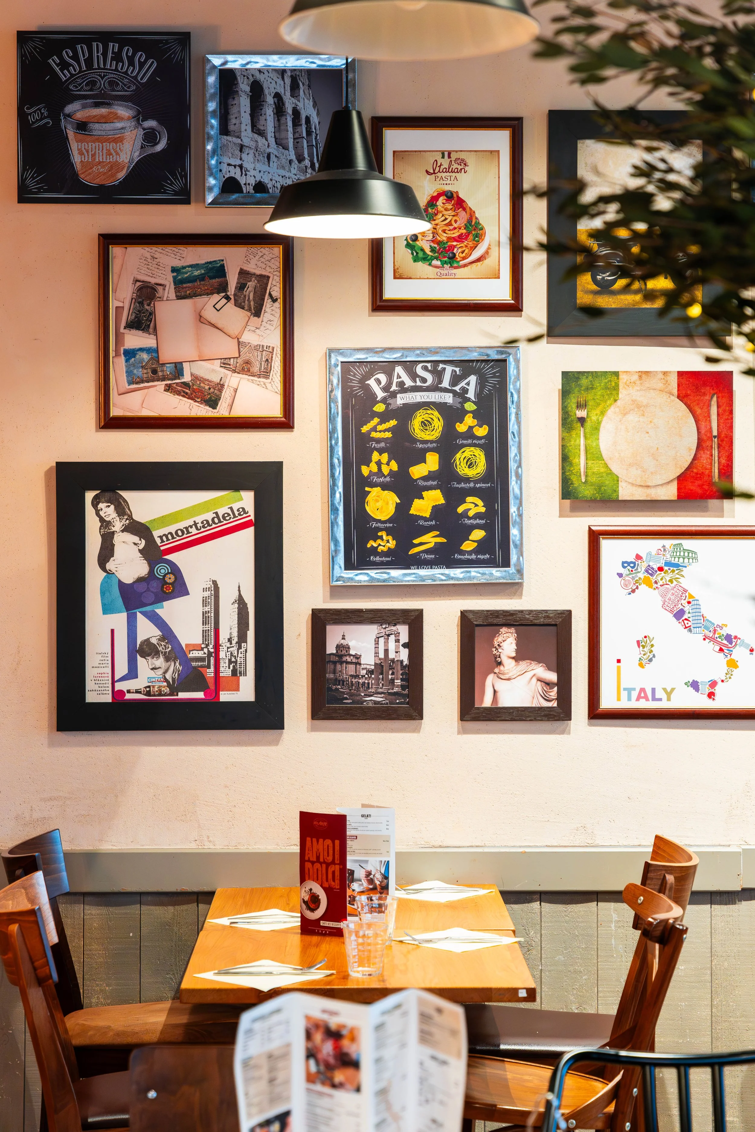 Une salle à manger de café ou restaurant avec des murs décorés de photos et illustrations, un éclairage suspendu, une table en bois avec des menus, des verres d'eau et des fourchettes et couteaux.
