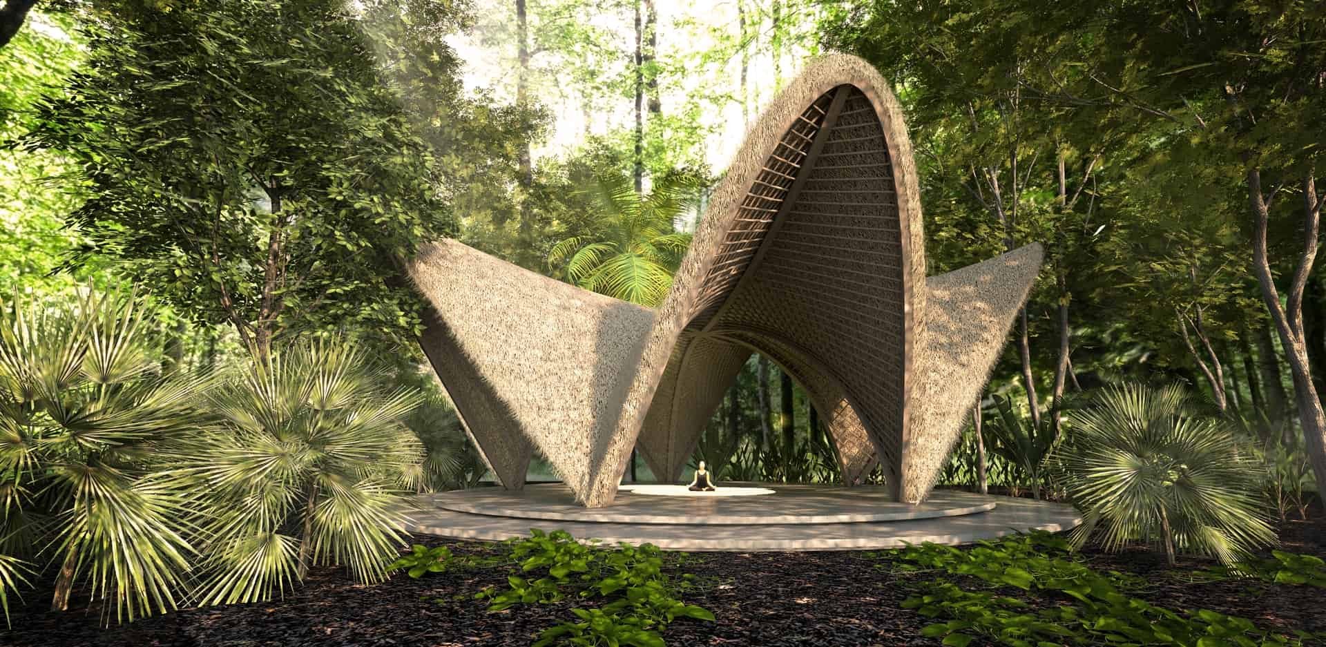 A modern pavilion with a thatched roof and open sides, situated in a lush, green forest environment. The pavilion is constructed with pointed, triangular shapes and features a platform where a person is sitting cross-legged.