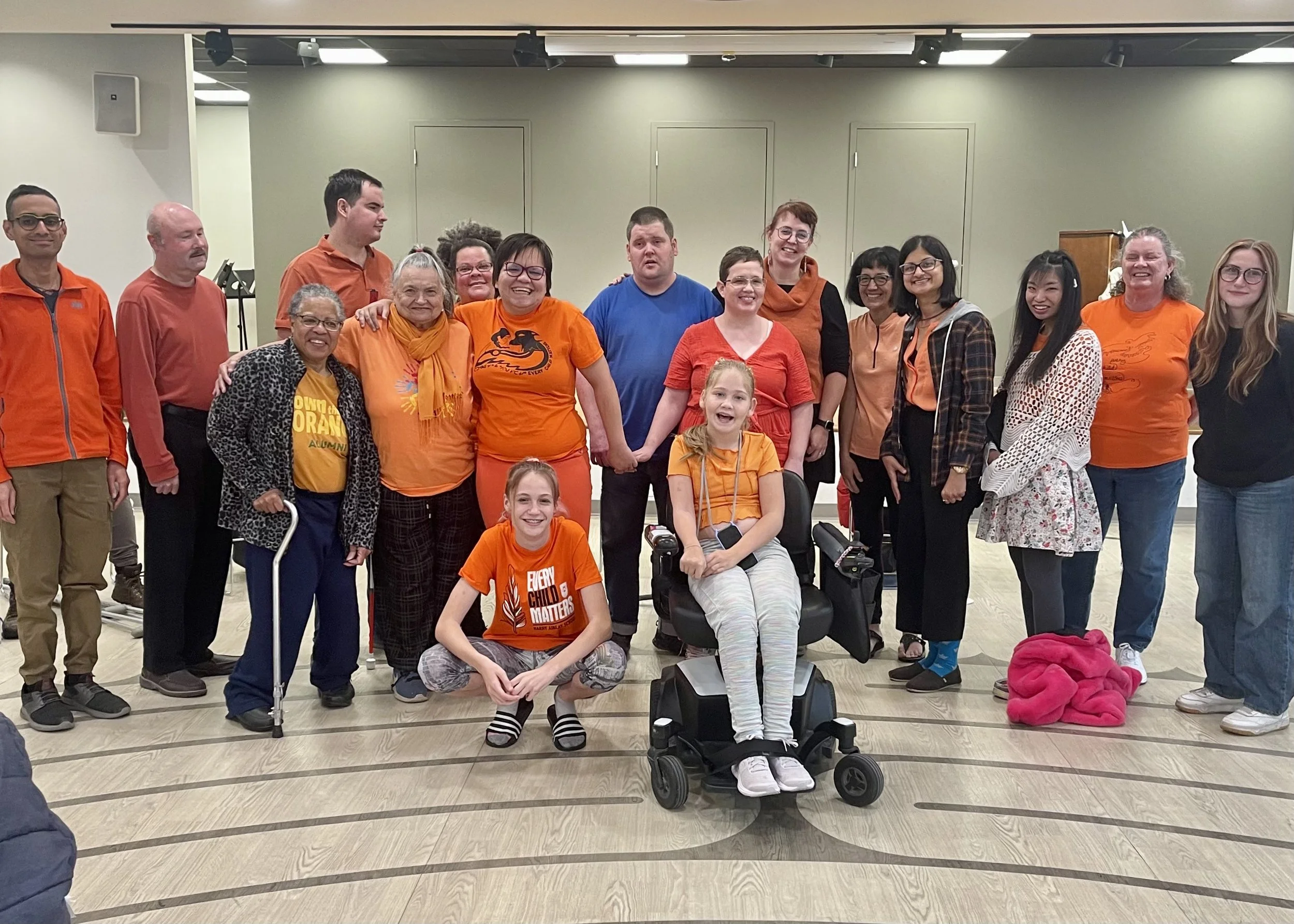 A diverse group of people of various ages, genders, and ethnicities are standing together indoors, some smiling and posing for the photo. They are dressed casually, with several individuals wearing orange clothing. There are young children, adults, and seniors in the group, and two of the children are seated in wheelchairs. The background features a plain wall with a closed door and some equipment, indicating the setting might be a community center or event hall.