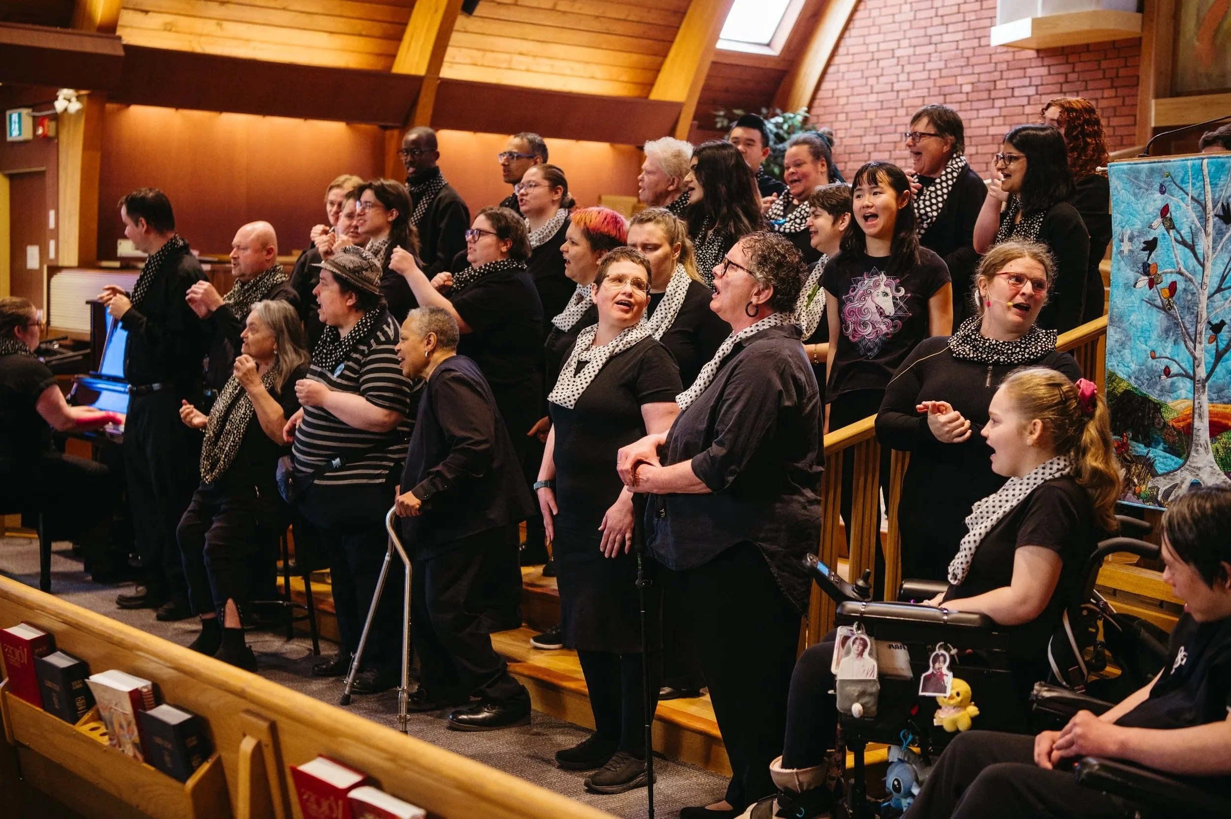 A diverse group of people, including children and adults, standing on wooden bleachers inside a church or community hall with wooden ceiling beams and brick walls, laughing and singing together.