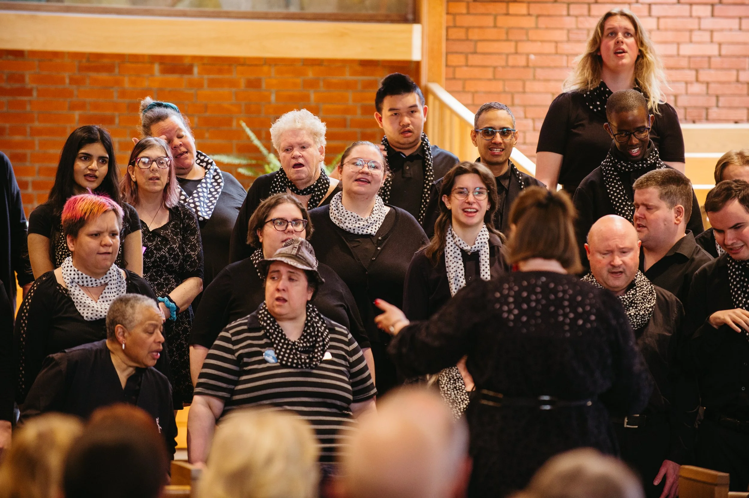 A diverse group of people singing together, wearing black clothing and polka dot accessories, in an indoor setting with brick walls.