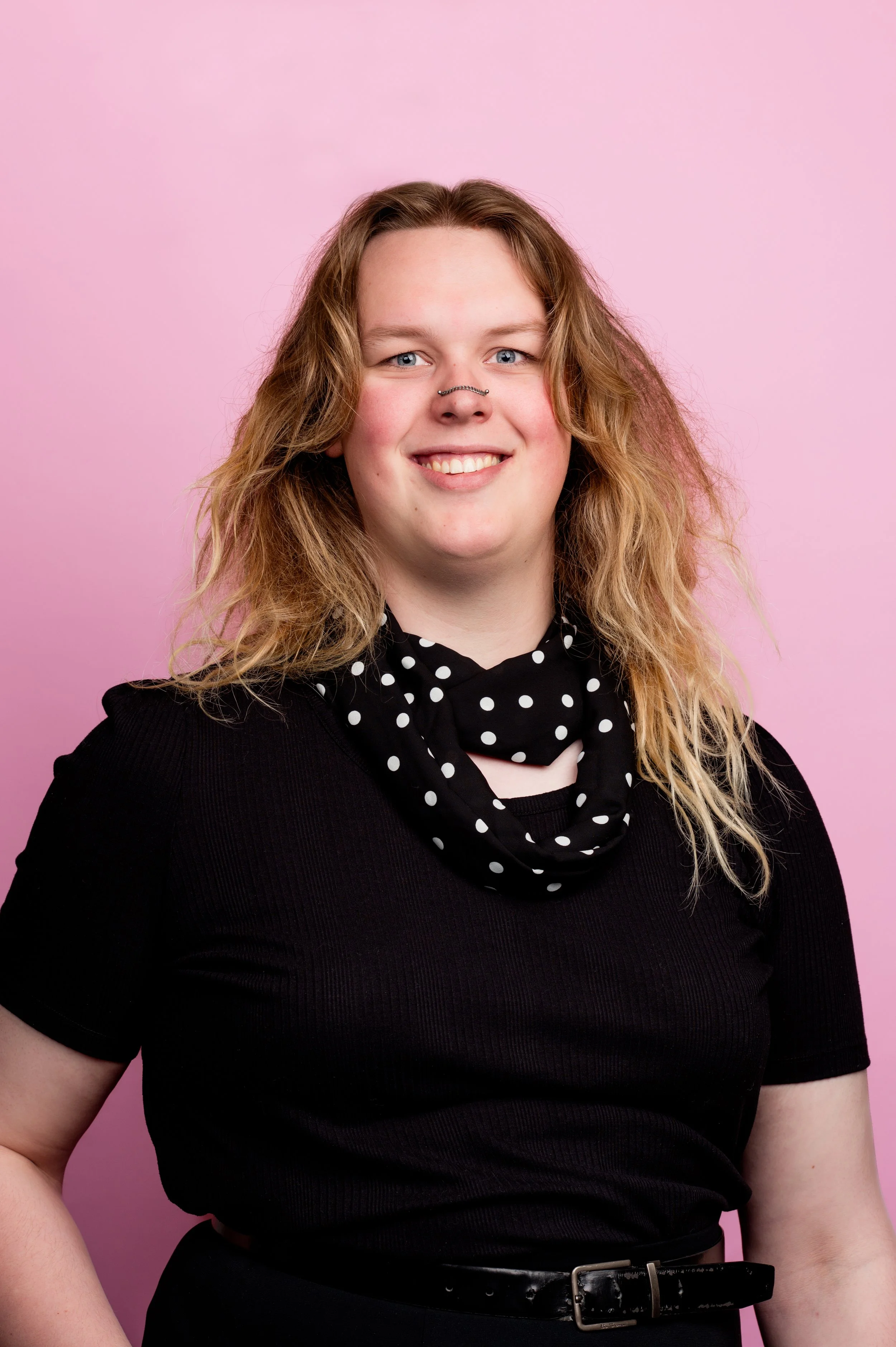 A woman with wavy light brown hair and blue eyes, smiling, wearing a black dress with a black and white polka dot neck scarf, standing against a pink background.