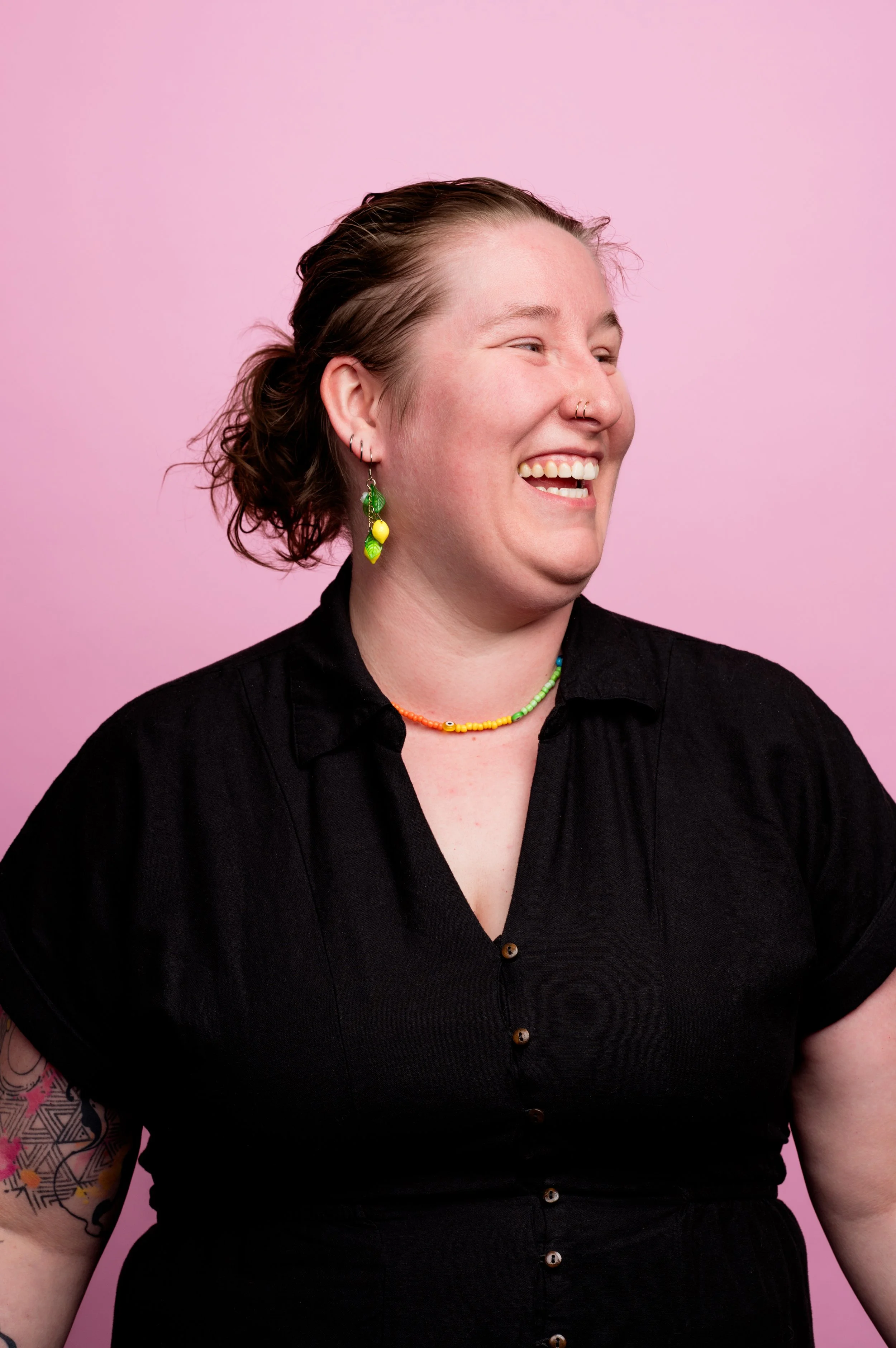 Close-up of a smiling person with short, wavy hair and colorful jewelry against a pink background.