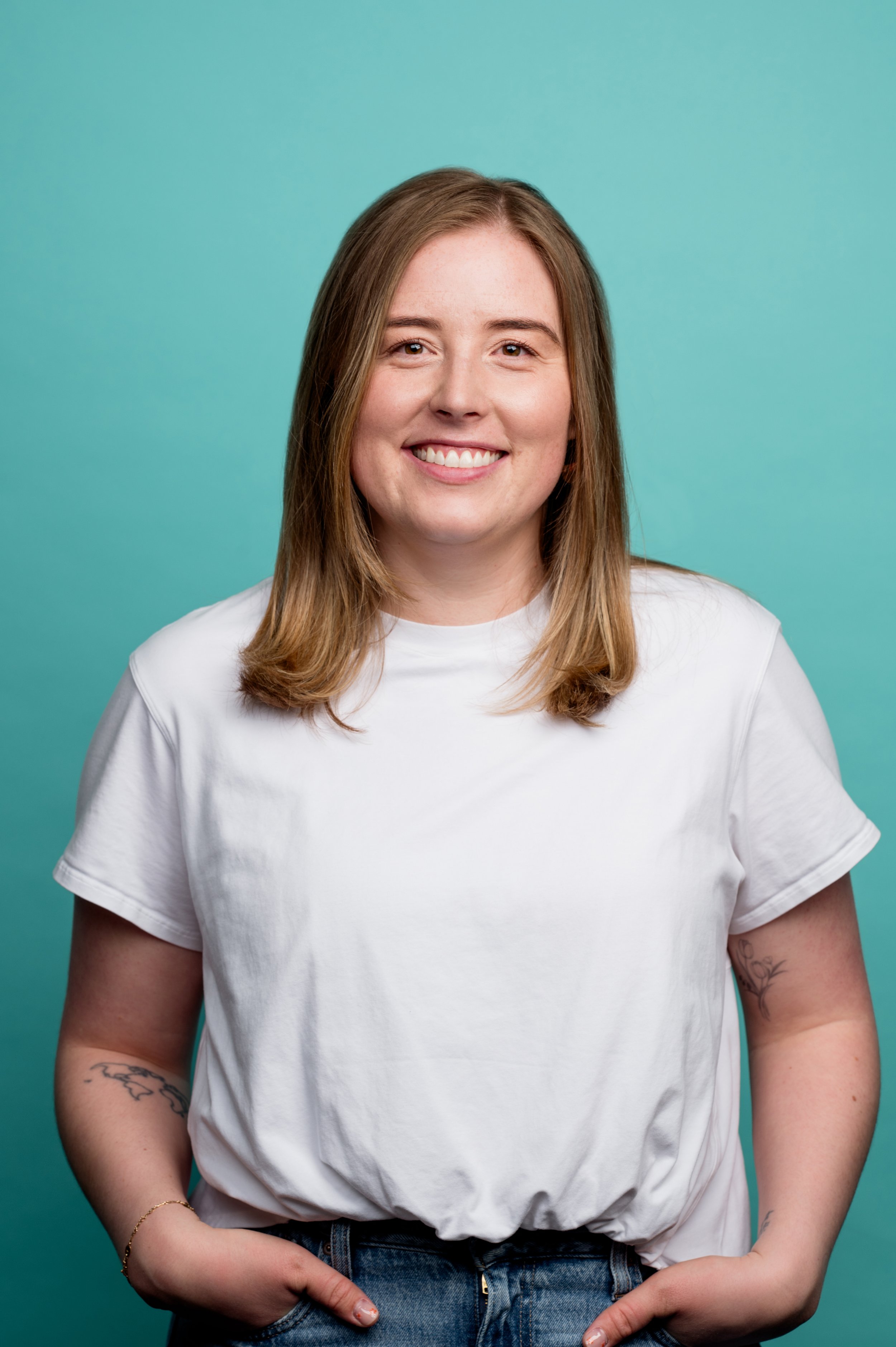 A young woman with light brown hair, smiling, wearing a white t-shirt, jeans, a gold bracelet, standing against a teal background.