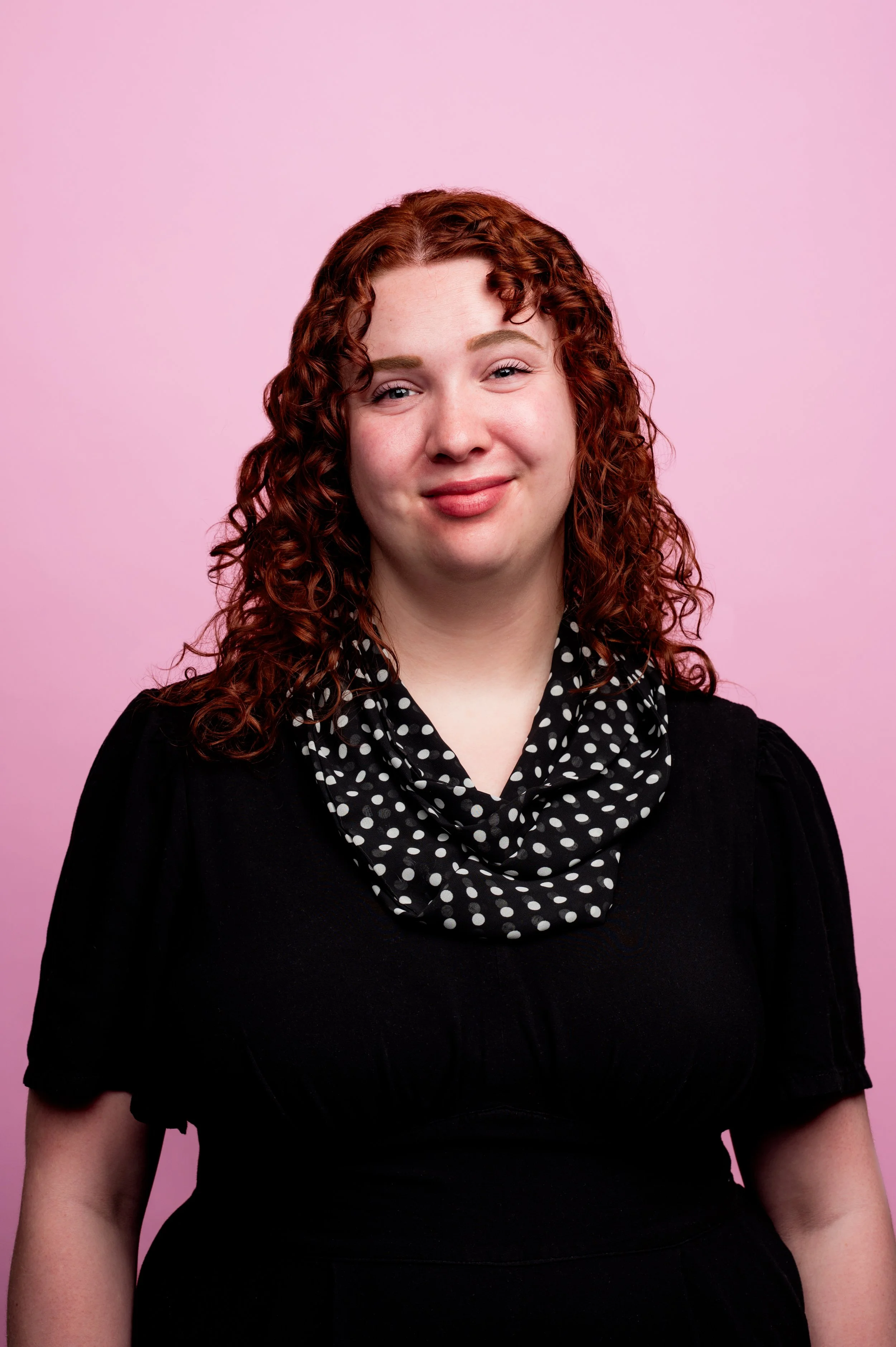 Portrait of a woman with curly red hair wearing a black dress and a polka dot scarf against a pink background.