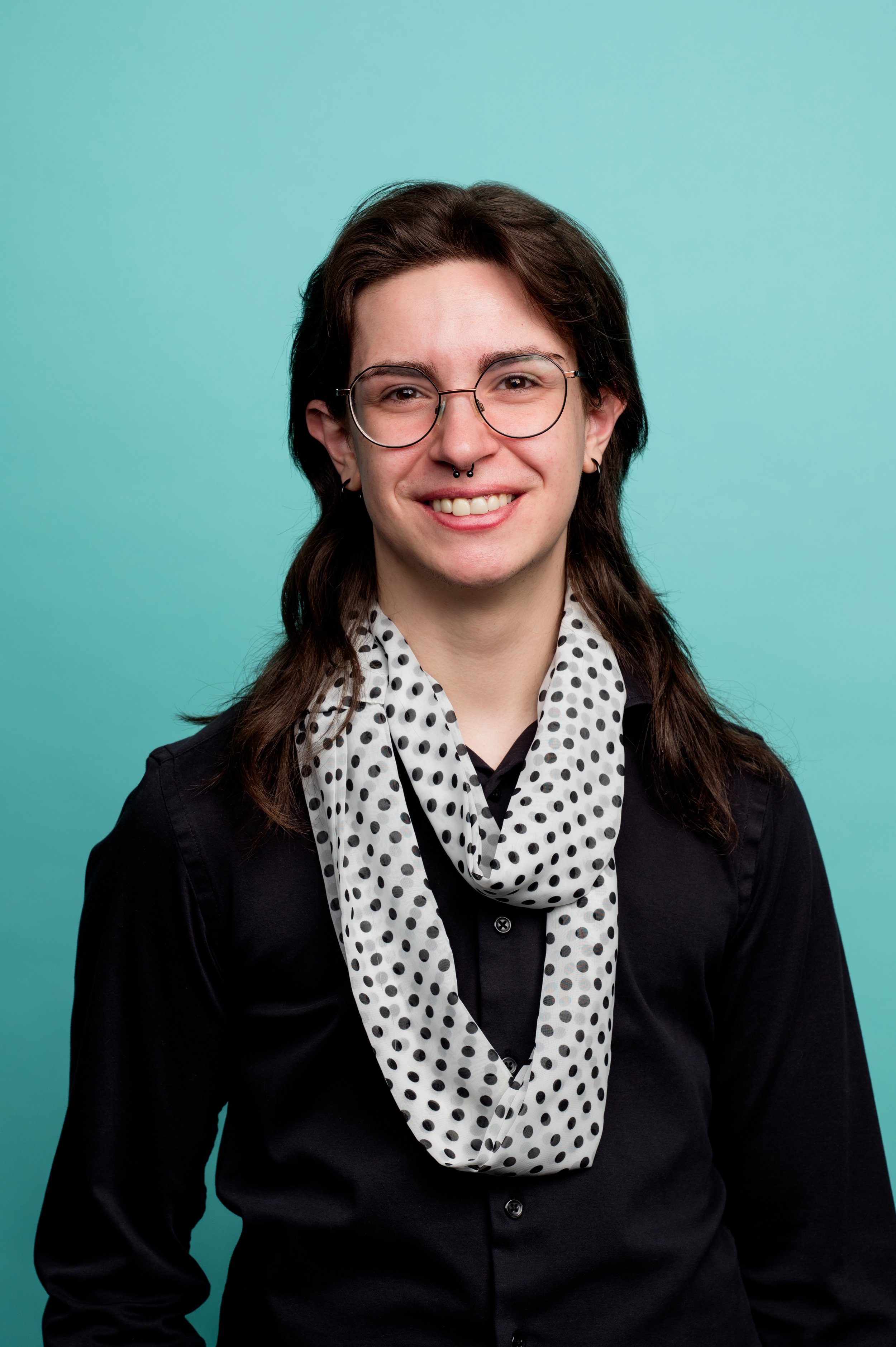 A young woman with glasses, dark hair, and a septum piercing, wearing a polka dot scarf, black shirt, and black blazer, standing against a turquoise background.