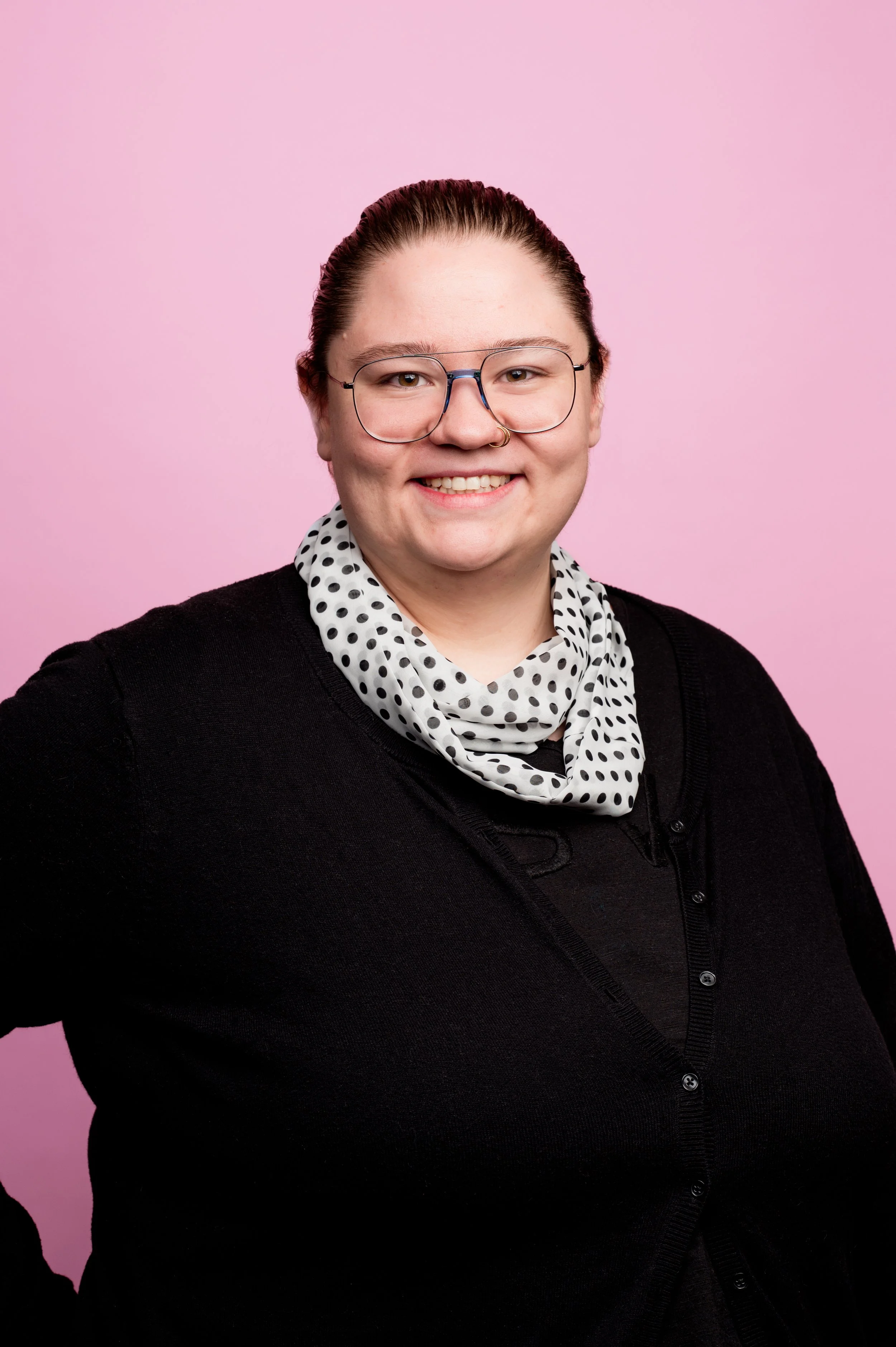 A person with glasses, a black sweater, a polka-dot scarf, and a nose ring, smiling in front of a pink background.