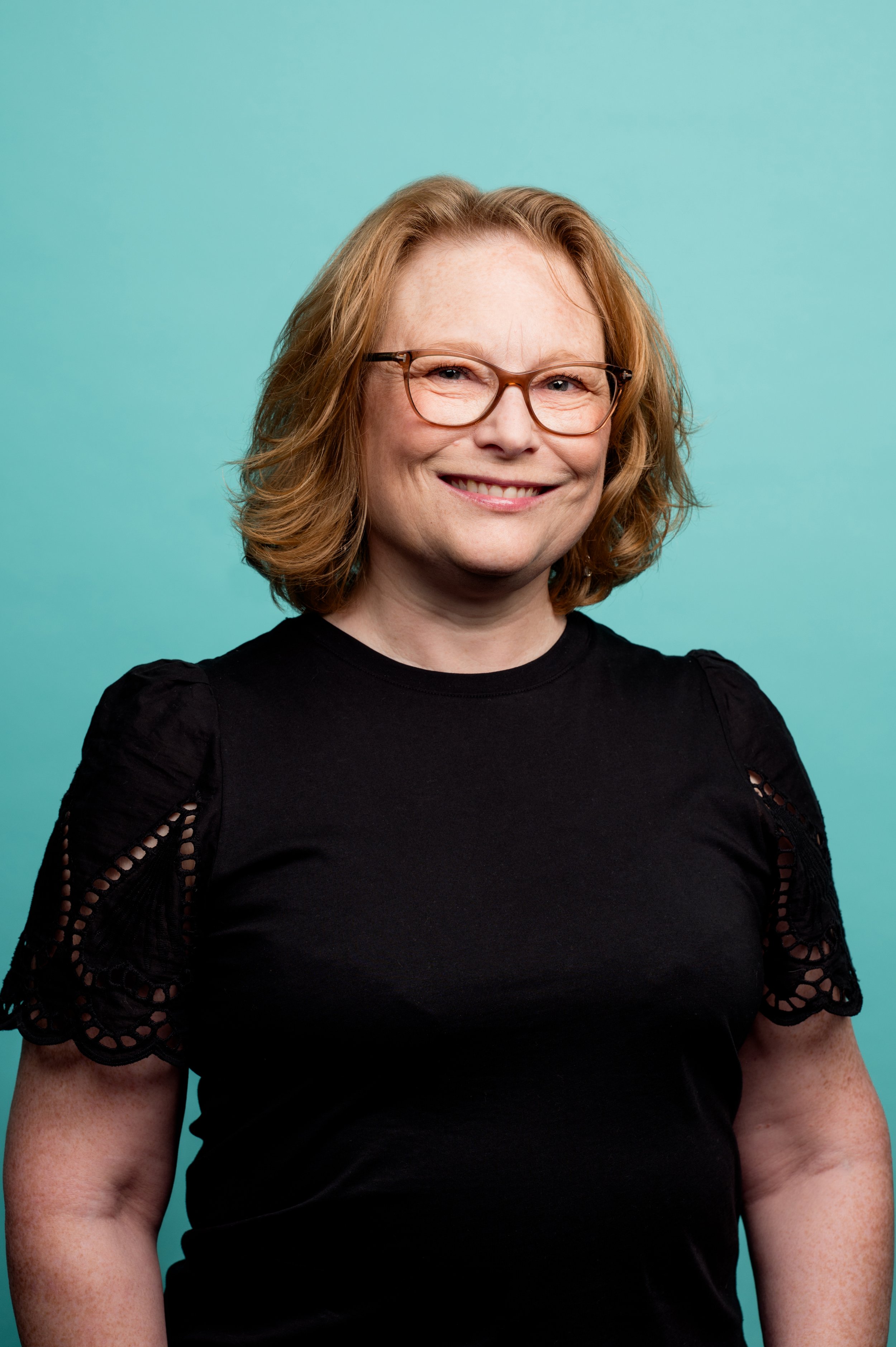 A woman with shoulder-length red hair, wearing glasses and a black top with lace sleeve details, smiling against a turquoise background.