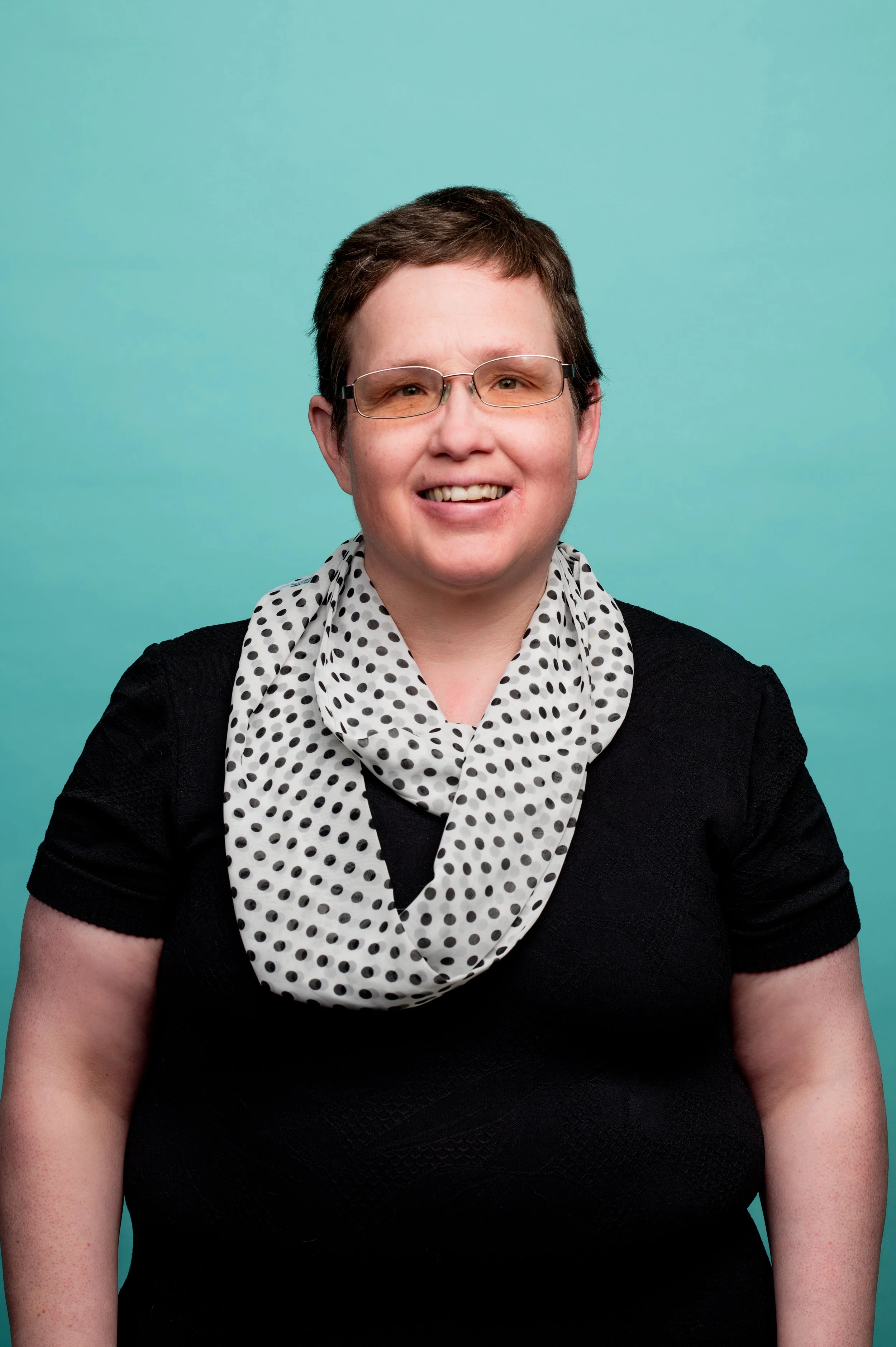 Close-up of a person with short brown hair, wearing glasses, a black top, and a white polka dot scarf, against a light blue background.
