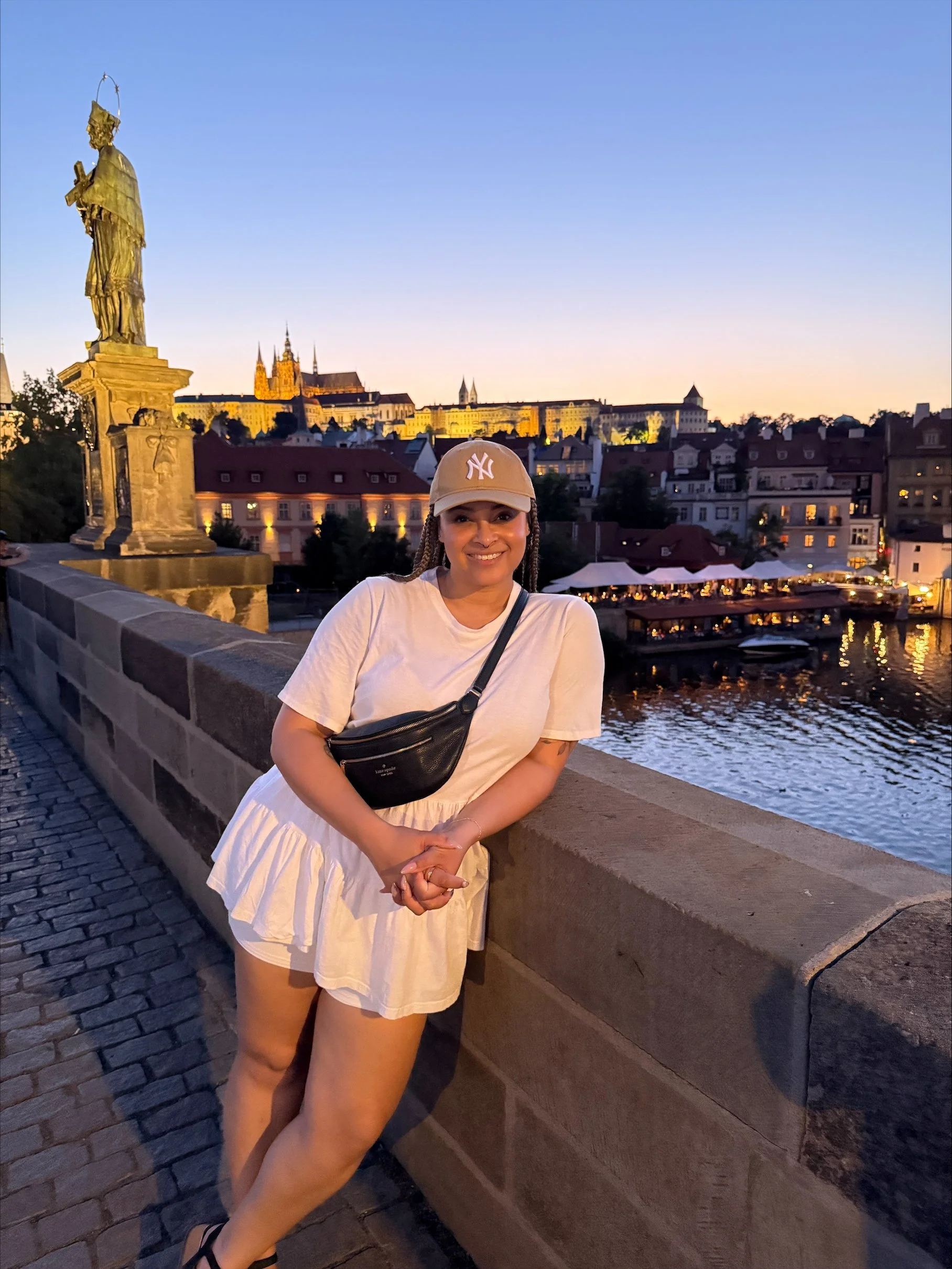 A woman wearing a beige cap, white shirt, and a black crossbody bag is smiling and posing by a stone railing on a riverbank, with city buildings and a castle in the background during sunset.