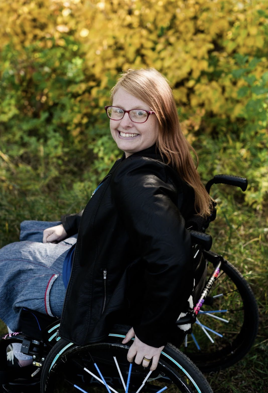 A smiling woman with red hair, glasses, and a black jacket sitting in a wheelchair outdoors with trees and yellow foliage in the background.