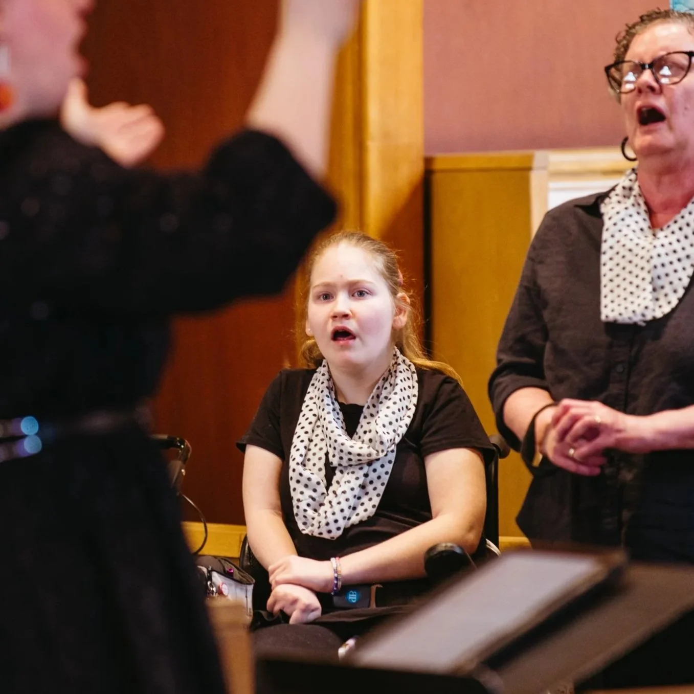 A young girl in a wheelchair with a polka dot scarf around her neck, looking surprised or confused, is sitting next to an older woman with matching polka dot scarf, both are in a room with wooden panels, and there is a blurred figure in black in the foreground.