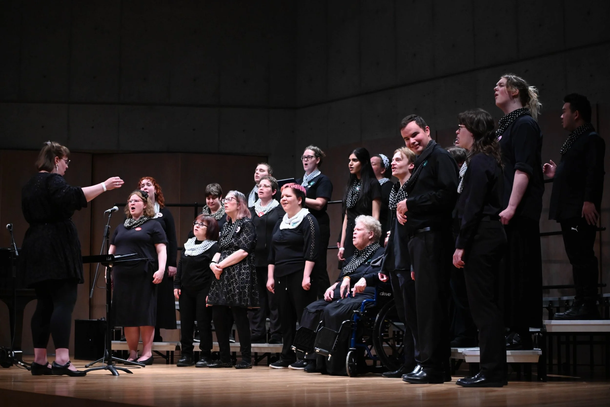 A choir of diverse individuals singing on stage guided by a conductor.