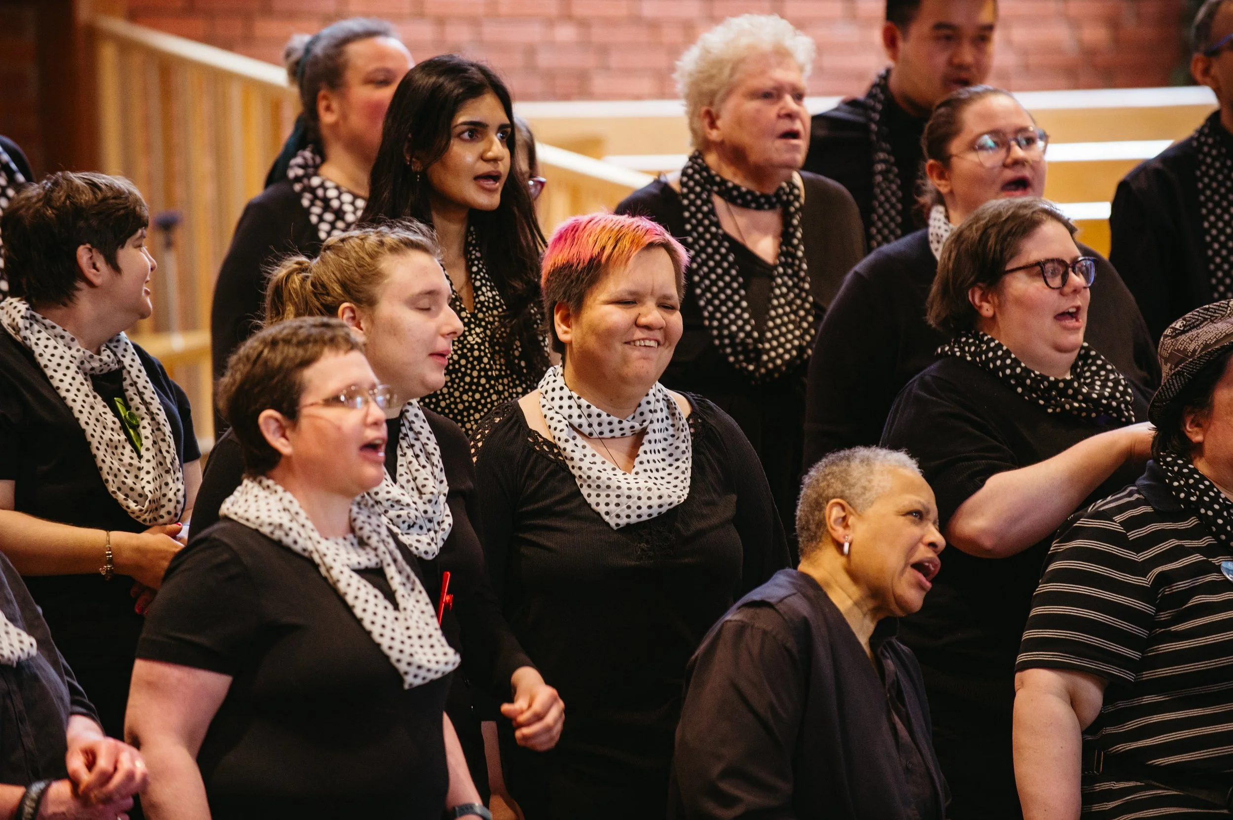 A diverse group of people singing together at a choir or concert, wearing black clothing with white polka-dotted scarves or accessories.