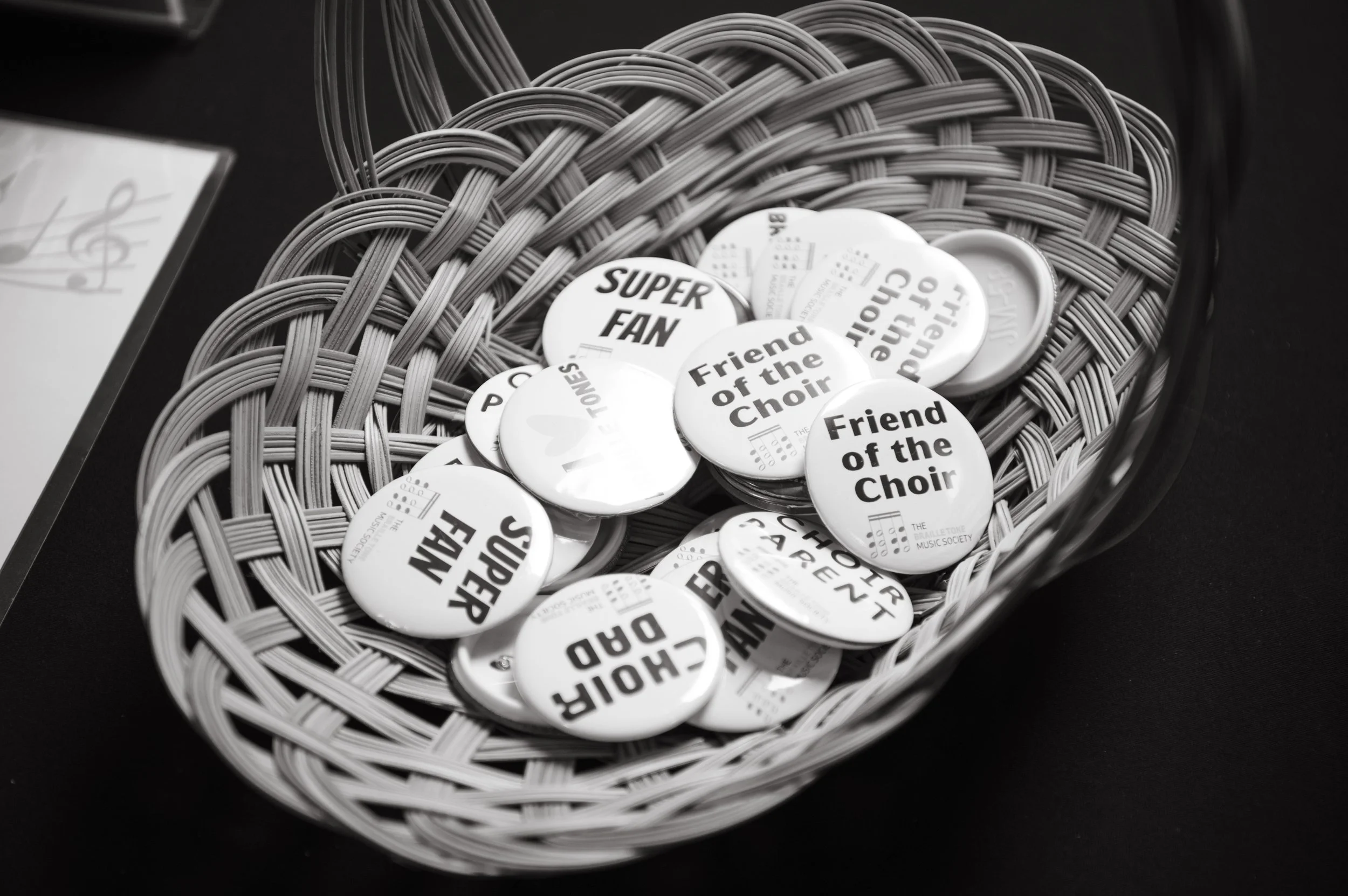 A basket containing round buttons with phrases like 'Friend of the Choir', 'Super Fan', and 'Proud Donor' printed on them, with some buttons showing musical notes and text from The Bullettone Music Society.