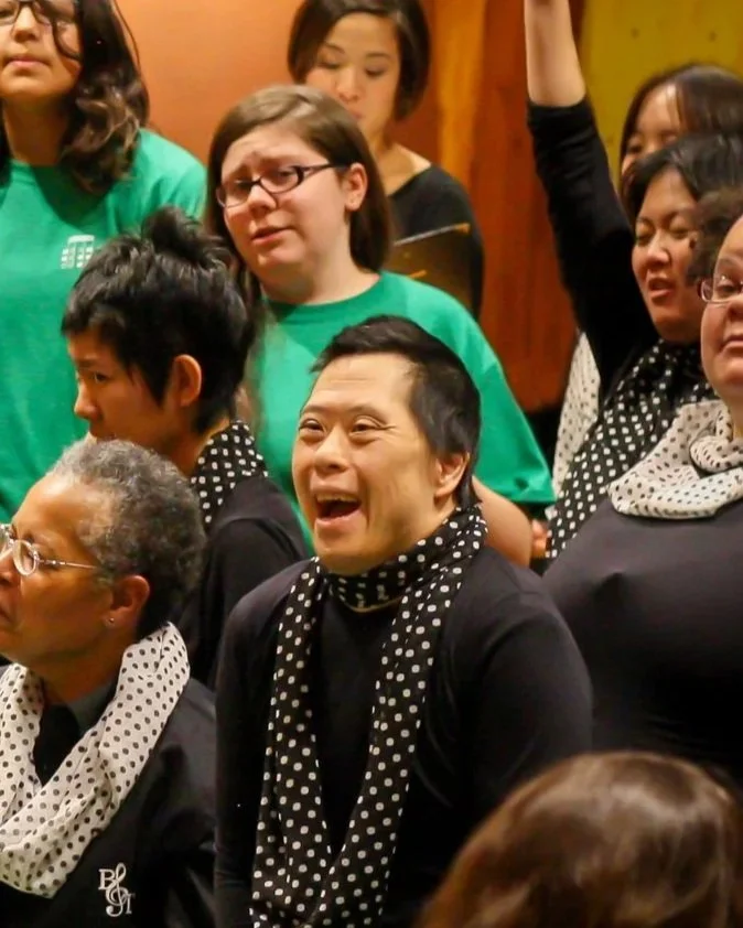 A group of diverse people, mostly women, gathered indoors. Some are smiling and laughing, with one woman raising her hand.