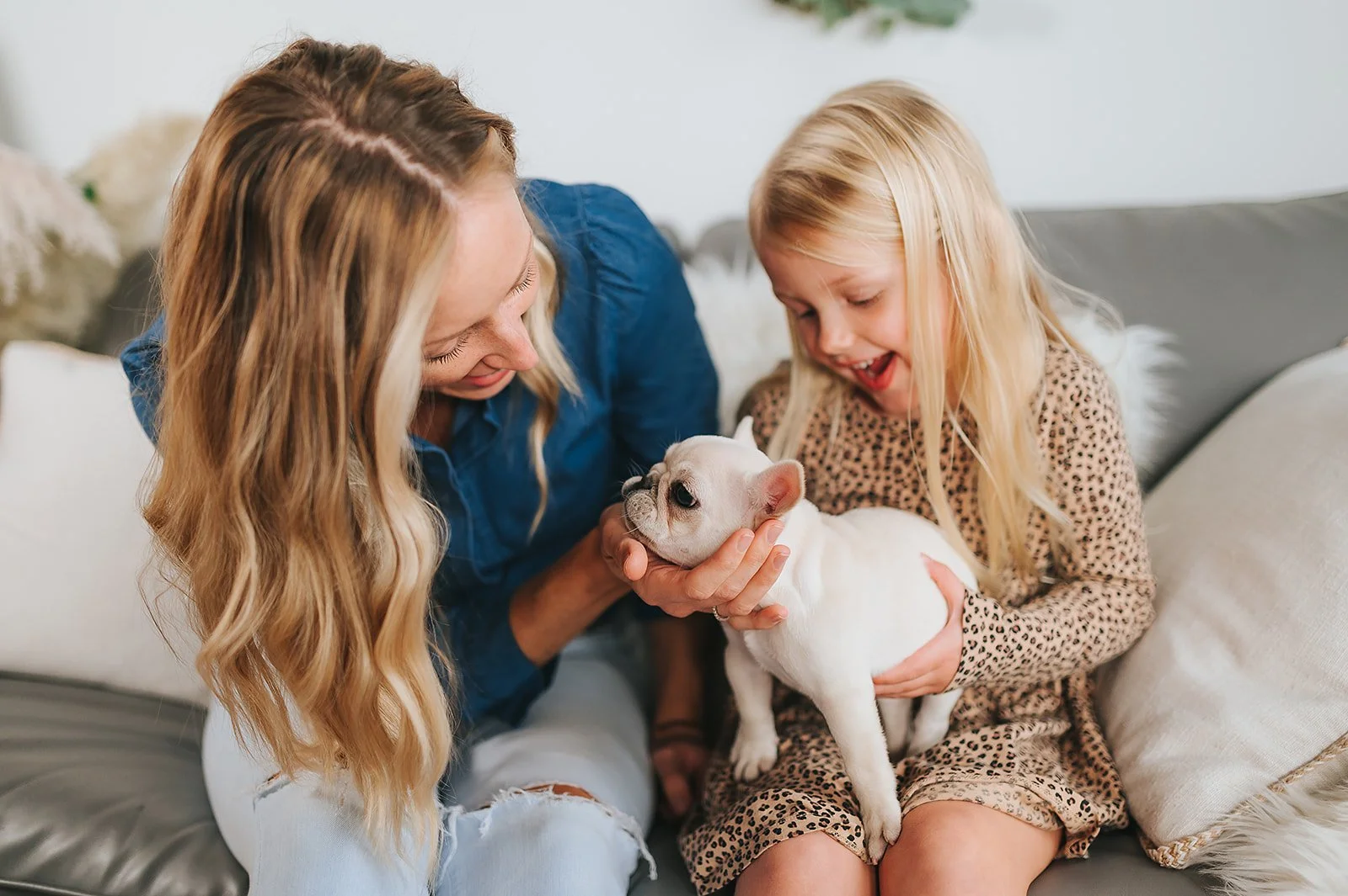 A woman and a young girl are happily playing with a small white cream French Bulldog puppy on a sofa in a cozy living room.