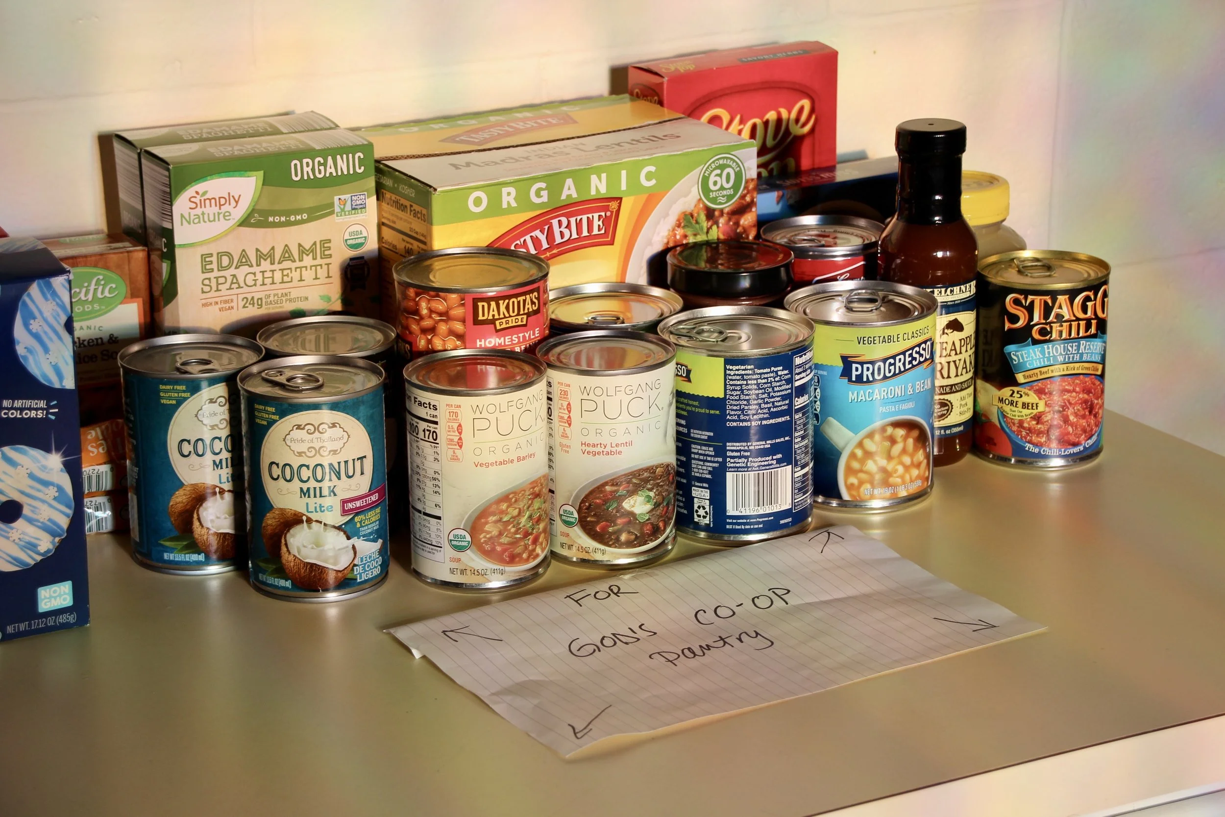 Shelf of canned foods, including various soups, beans, coconut milk, and chili, with a handwritten note on paper in front.