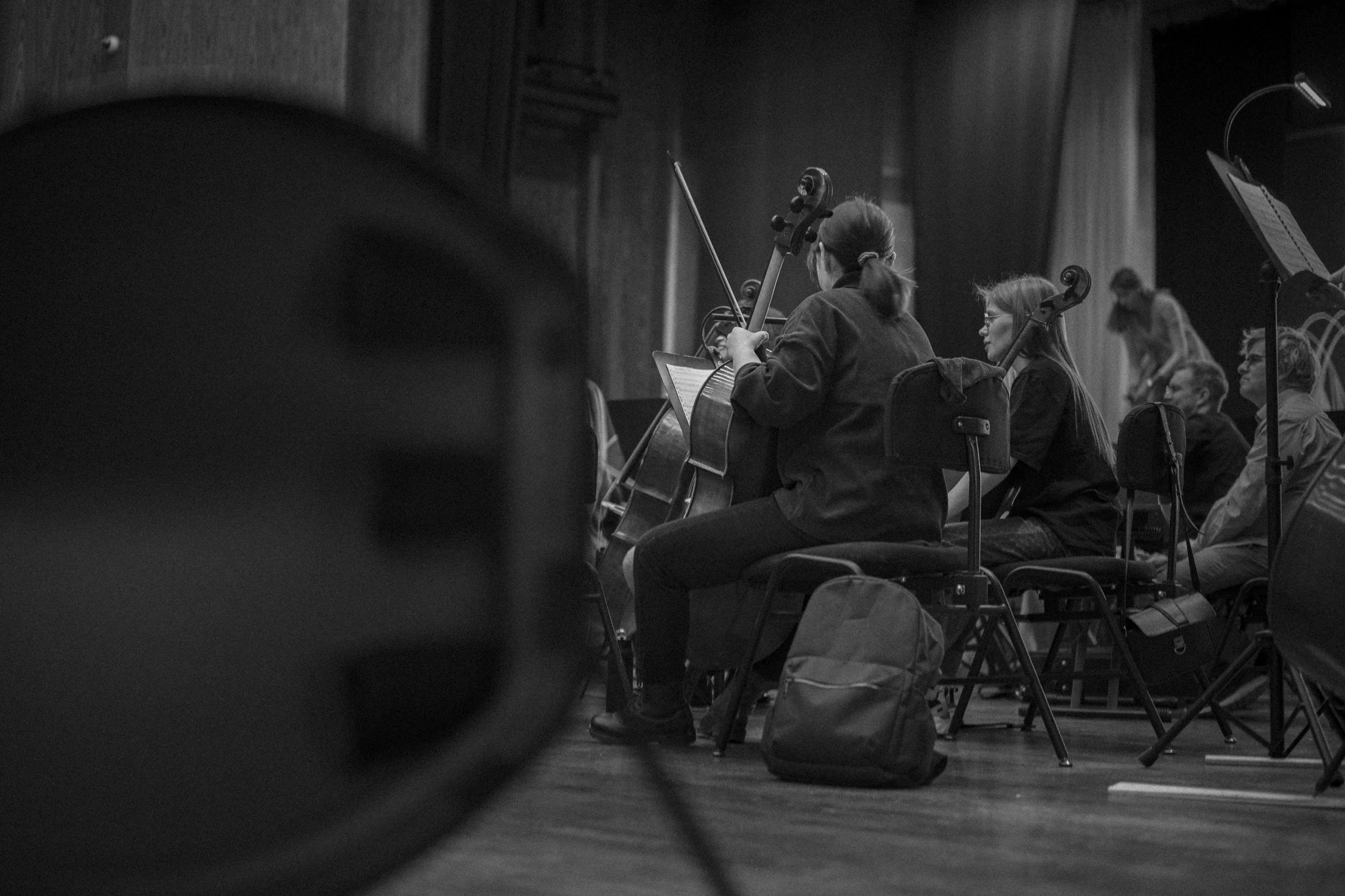 Black-and-white photograph of musicians sitting on chairs, holding string instruments such as violins, rehearsing or performing in a hall with curtains and sheet music stands.