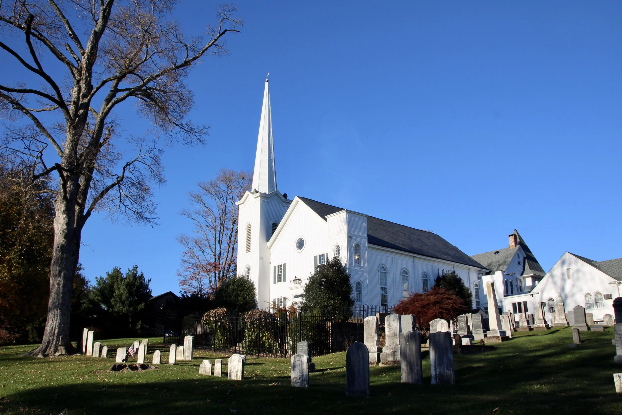 A white church with a tall steeple surrounded by leafless trees and a graveyard with headstones, under a clear blue sky.
