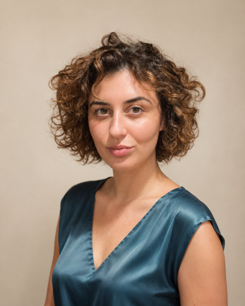 A woman with short, curly brown hair wearing a blue satin sleeveless top, looking at the camera with a neutral expression against a plain beige background.