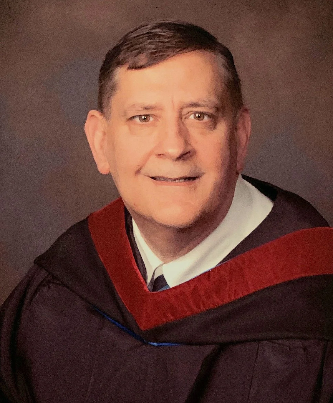 A senior man in academic regalia, wearing a black gown with a red velvet panel and white shirt, posing for a portrait.