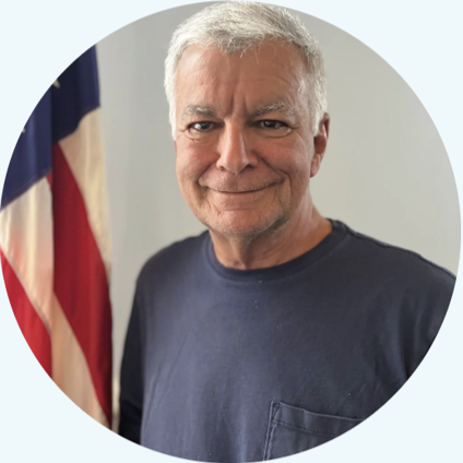 An older man with gray hair wearing a navy blue t-shirt, standing in front of an American flag.