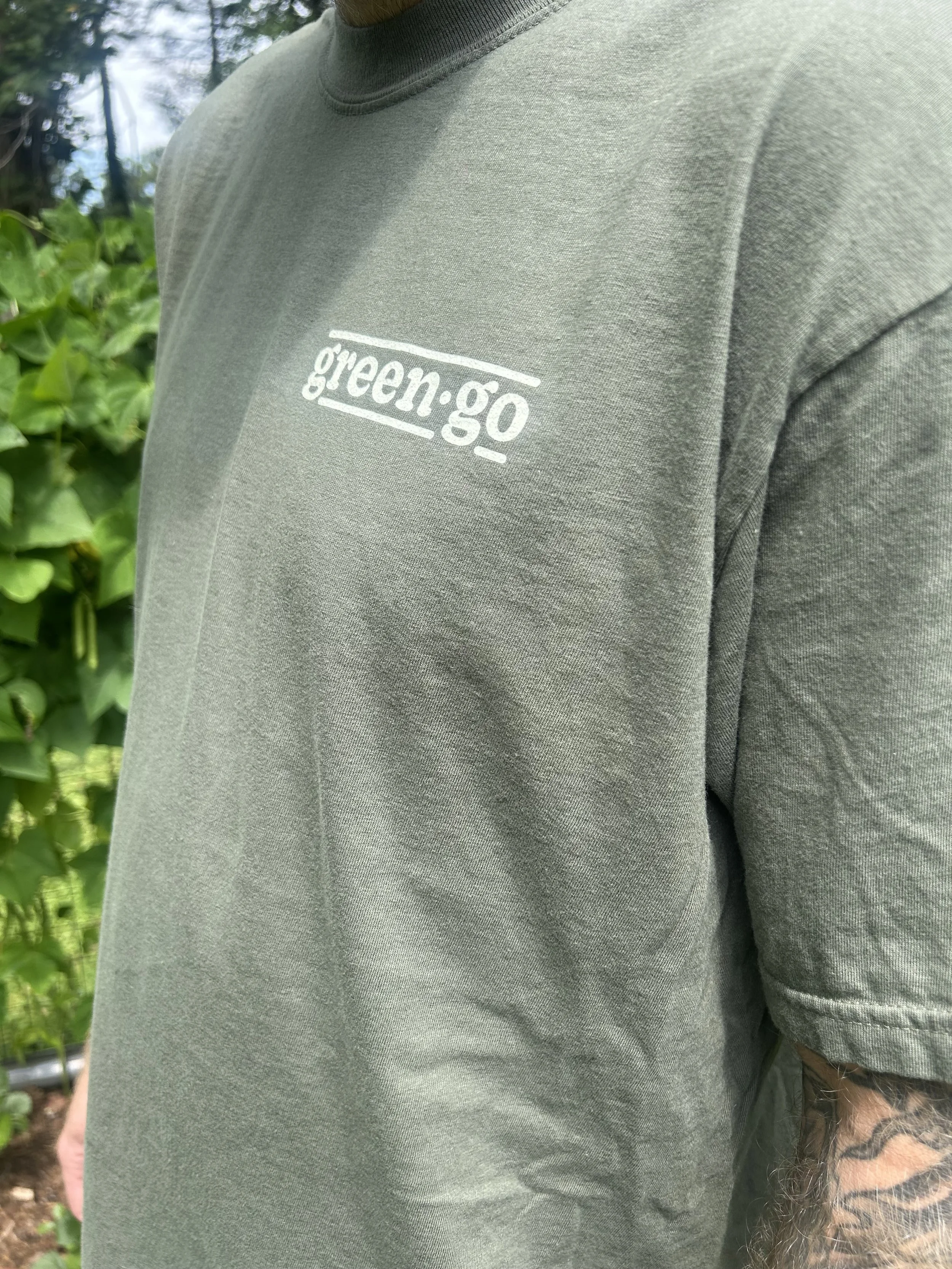 Front chest close-up of army green Green Go Cleaning Co t-shirt showing green·go wordmark logo cream screen print on tattooed man in Pisgah Forest NC garden woman-owned cleaning brand merch