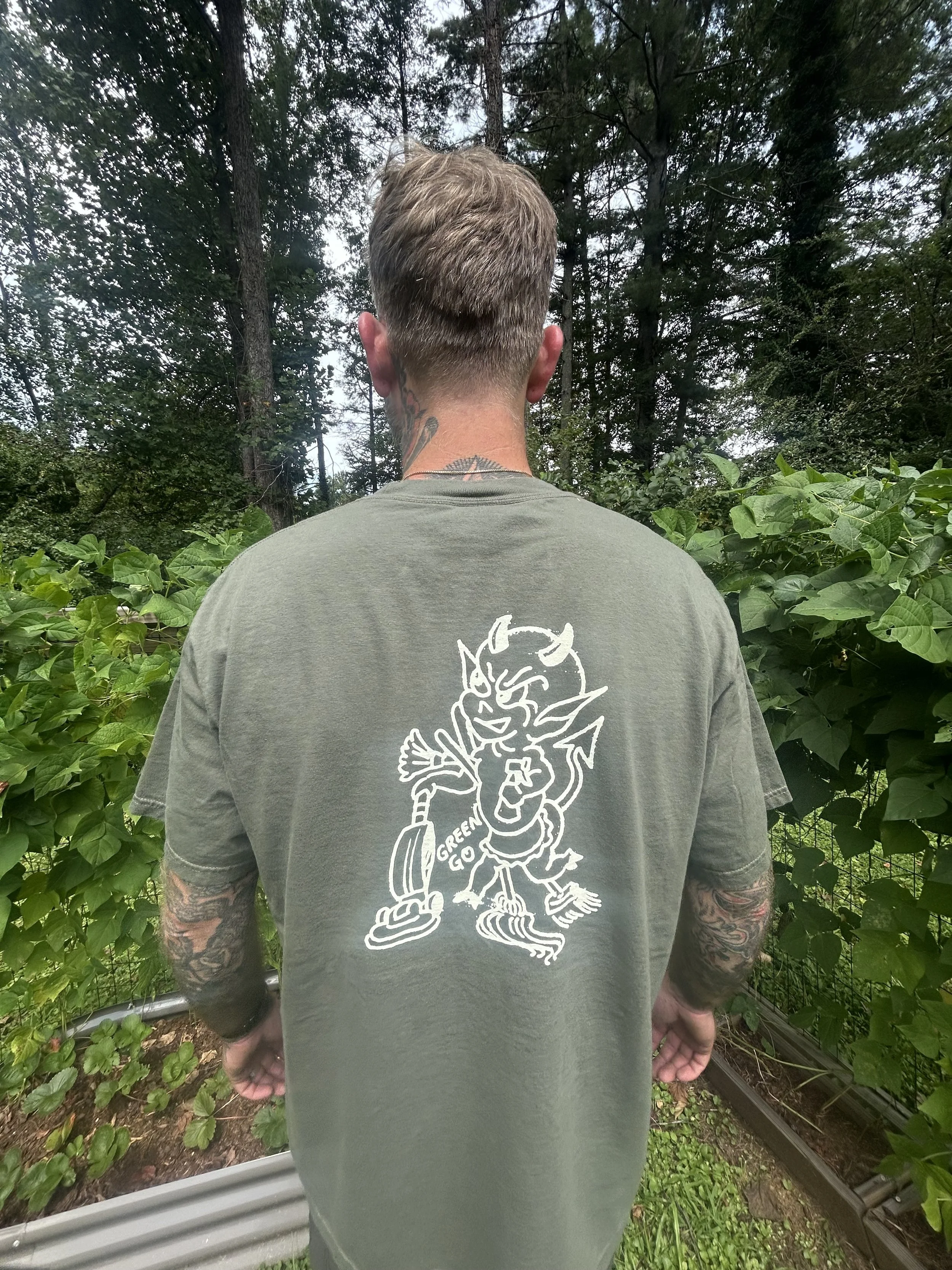 Back of army green Green Go Cleaning Co t-shirt showing vintage cartoon devil with cleaning supplies screen printed in cream on tattooed man standing in forest Pisgah Forest NC small batch merch
