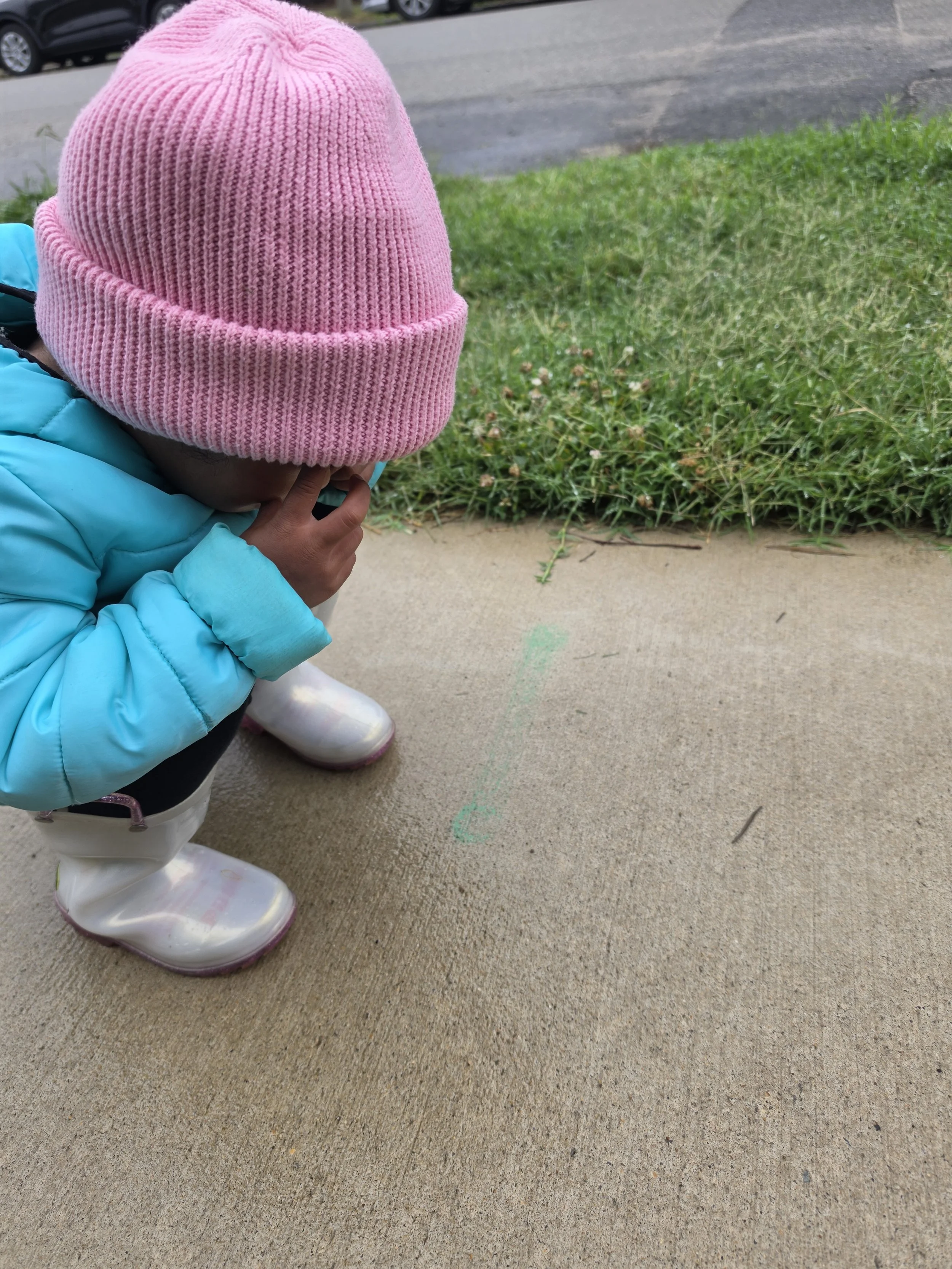 A toddler in a pink hat and blue coat has their fingers near their eyes like make believe binoculars and is looking at a worm.