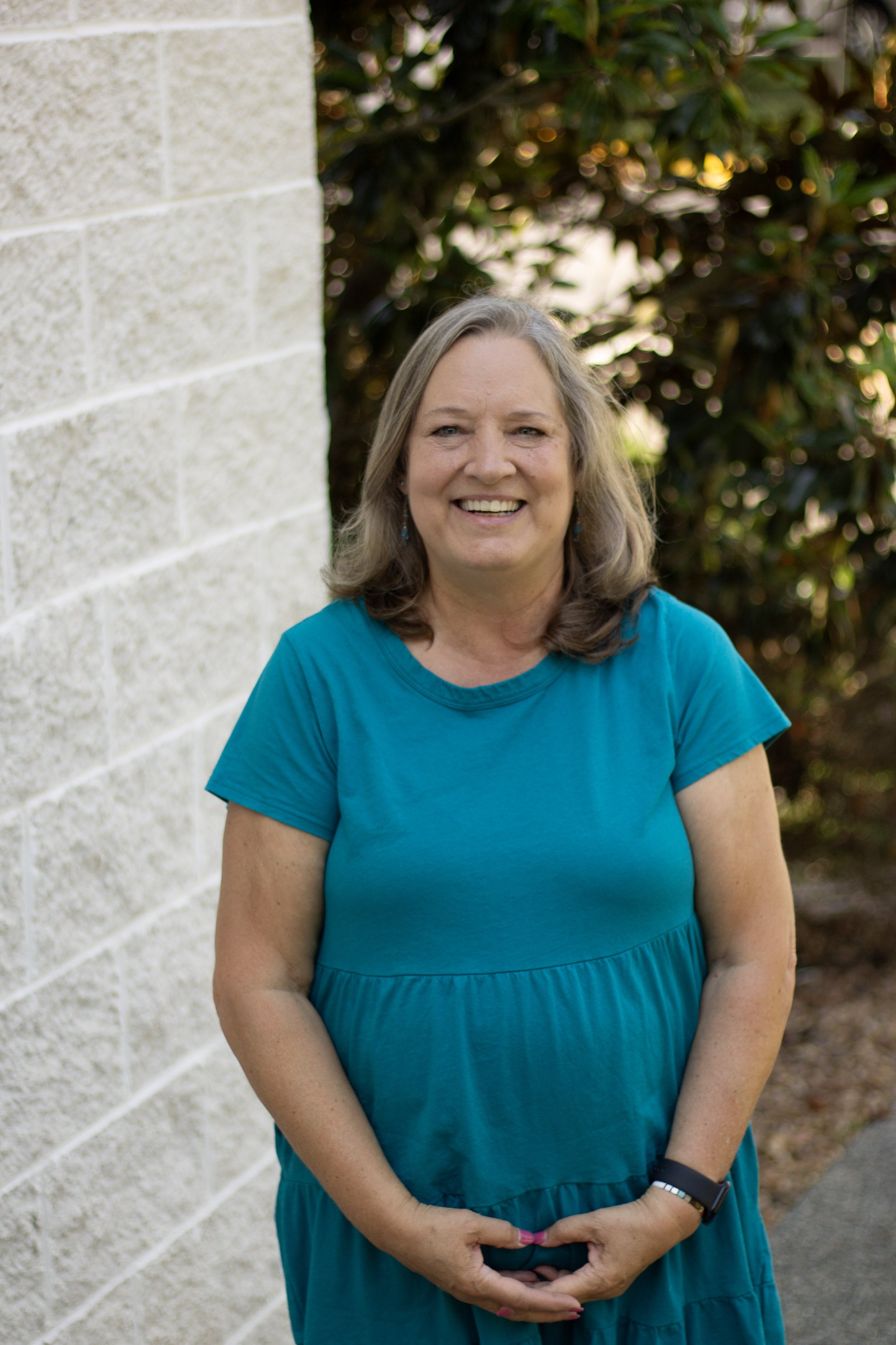 Woman with brown hair, wearing a blue short-sleeve button-up shirt and beige pants, standing outdoors beside a white brick wall with green bushes in the background.