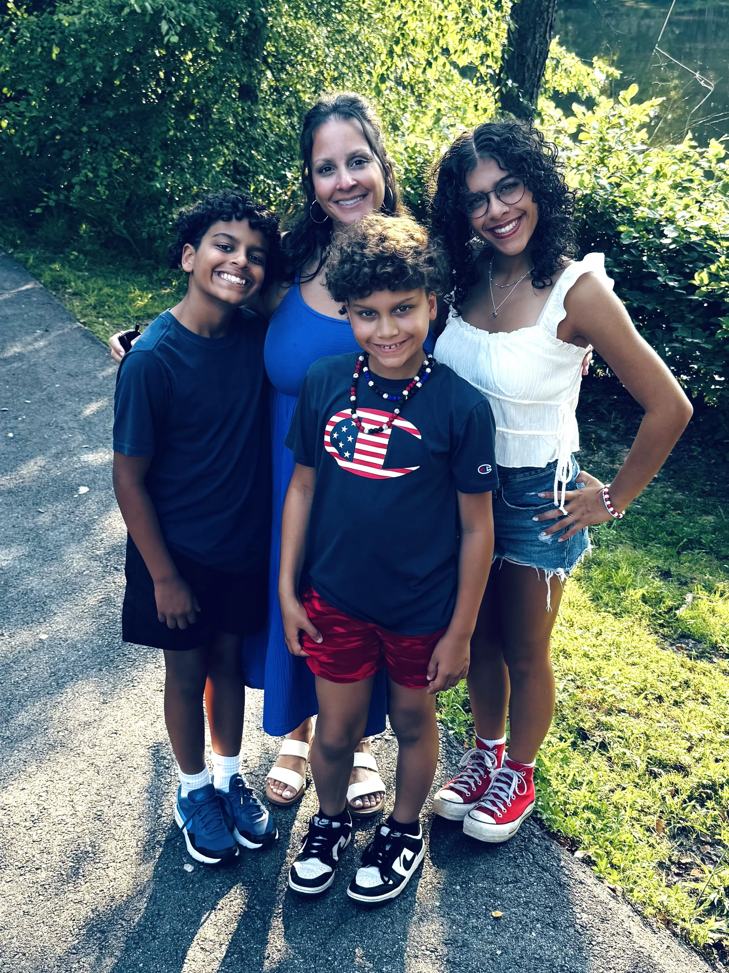 A group of four smiling people, two women and two boys, standing outdoors on a shaded sidewalk with greenery in the background. At a private Christian school in Chesterfield and Midlothian