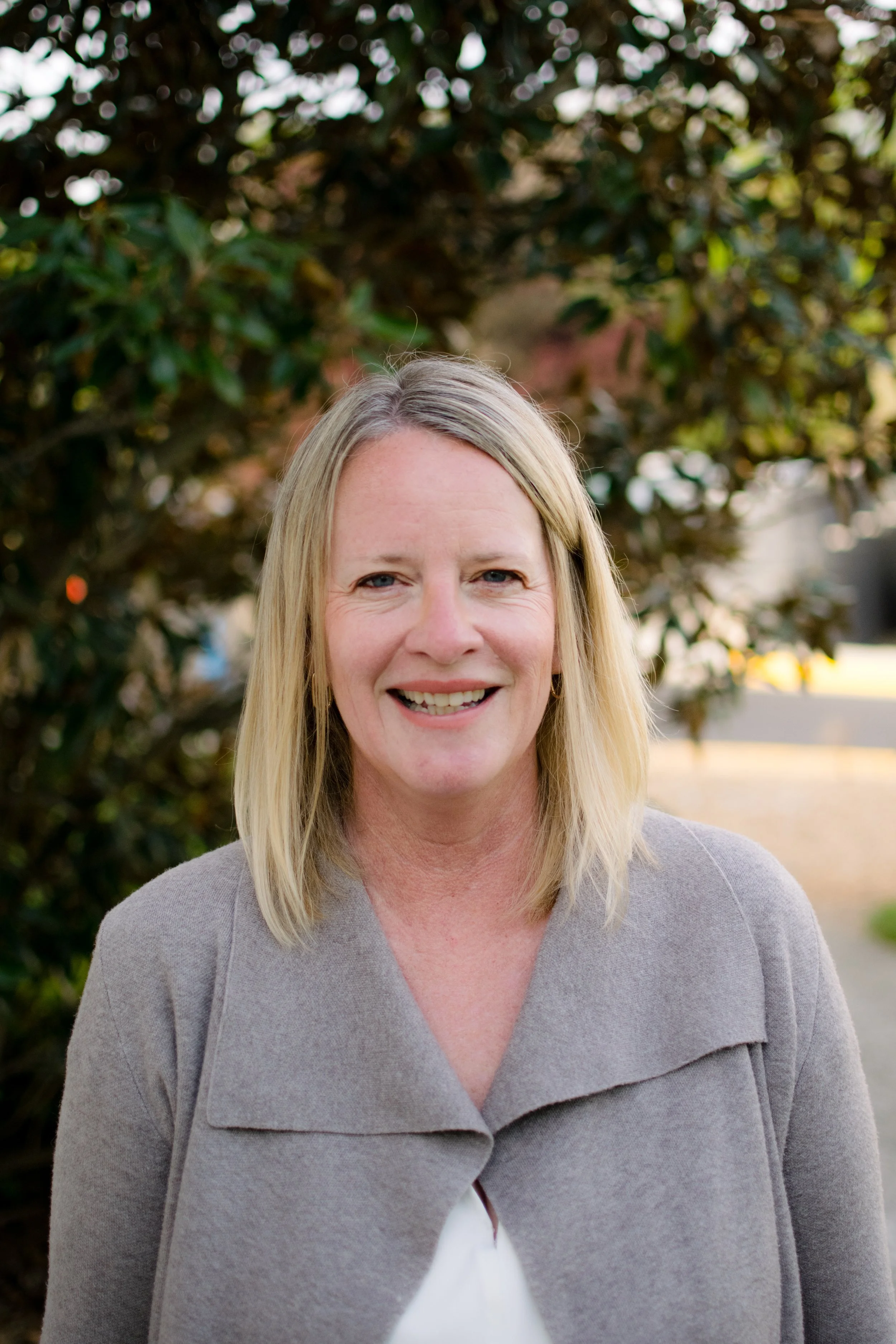 A smiling middle-aged woman with blonde hair, wearing a denim jacket and a white shirt with black horizontal stripes, standing in a hallway with blurred background.