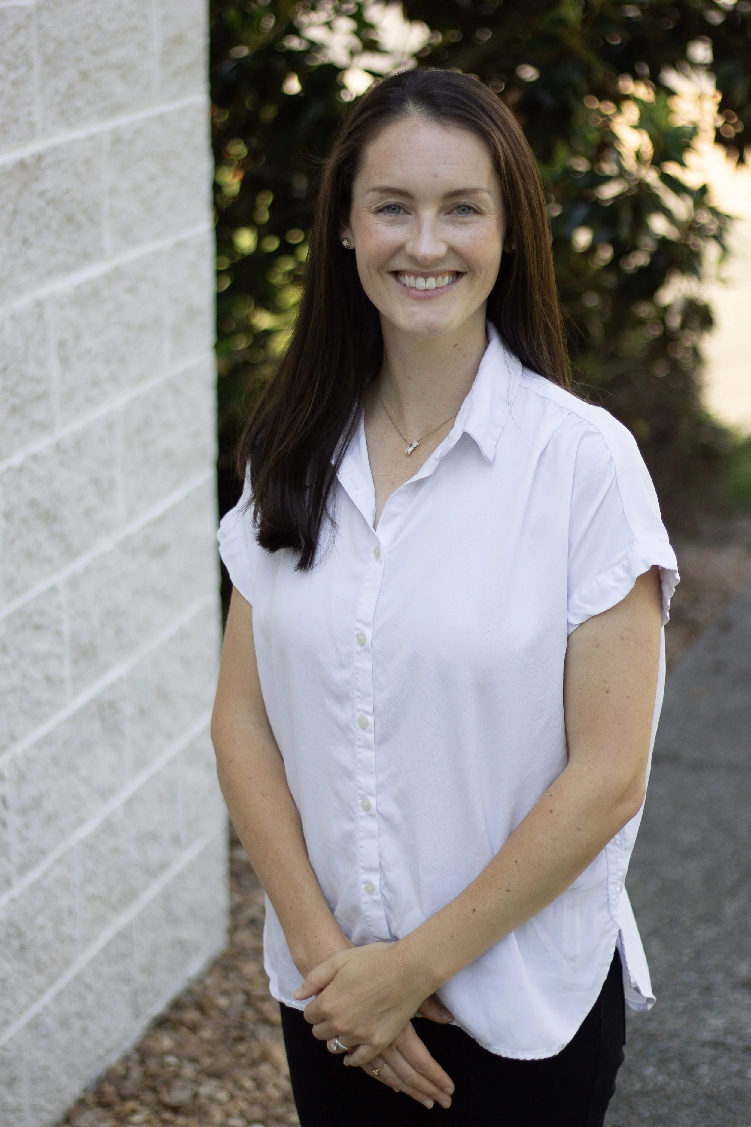 A woman with long dark hair smiling, wearing a white shirt and standing outdoors next to a light-colored brick wall with greenery in the background.