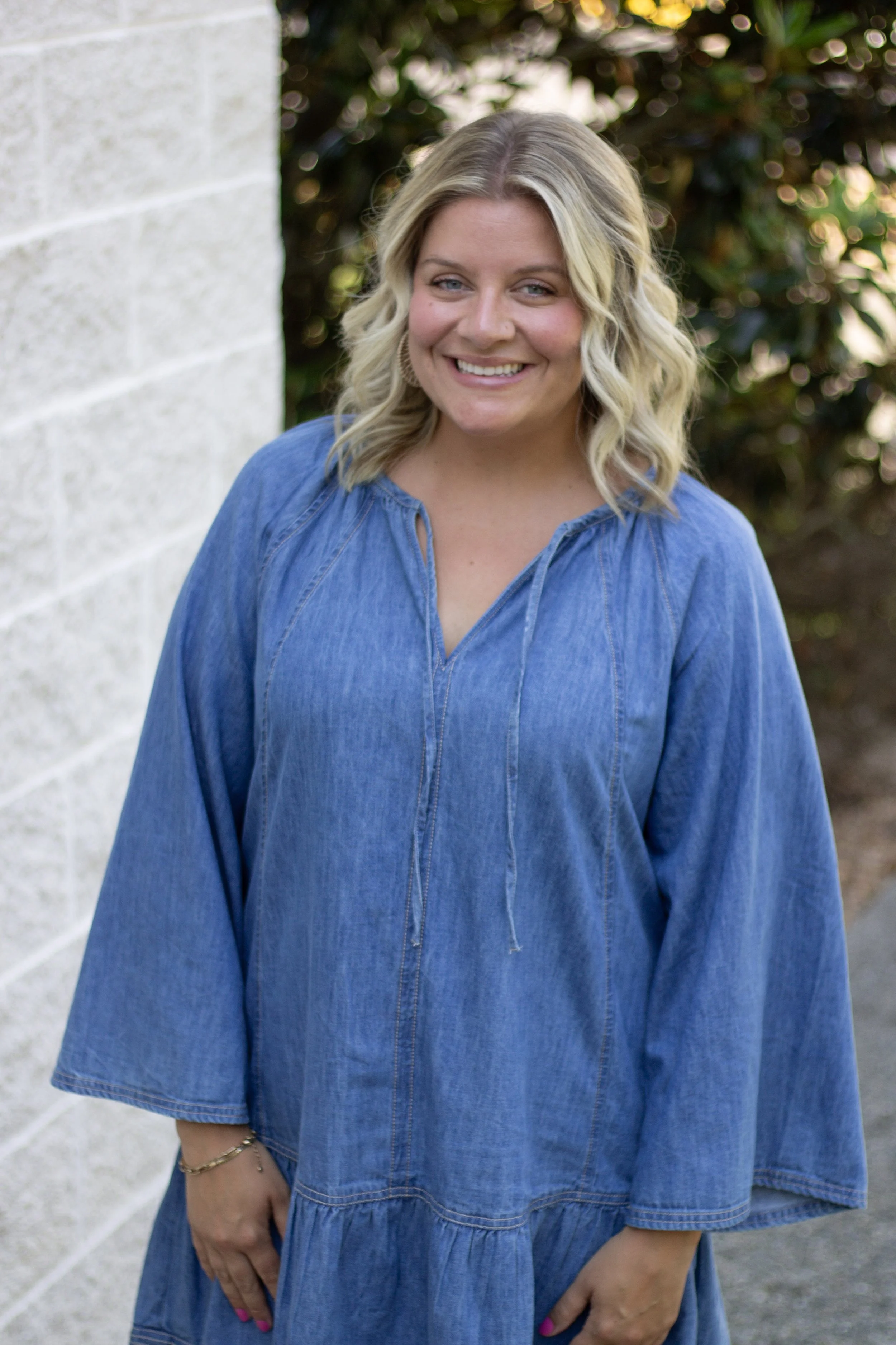 A woman with blonde, wavy hair dressed in a loose-fitting blue denim dress, smiling outdoors near a white brick wall with green foliage in the background.