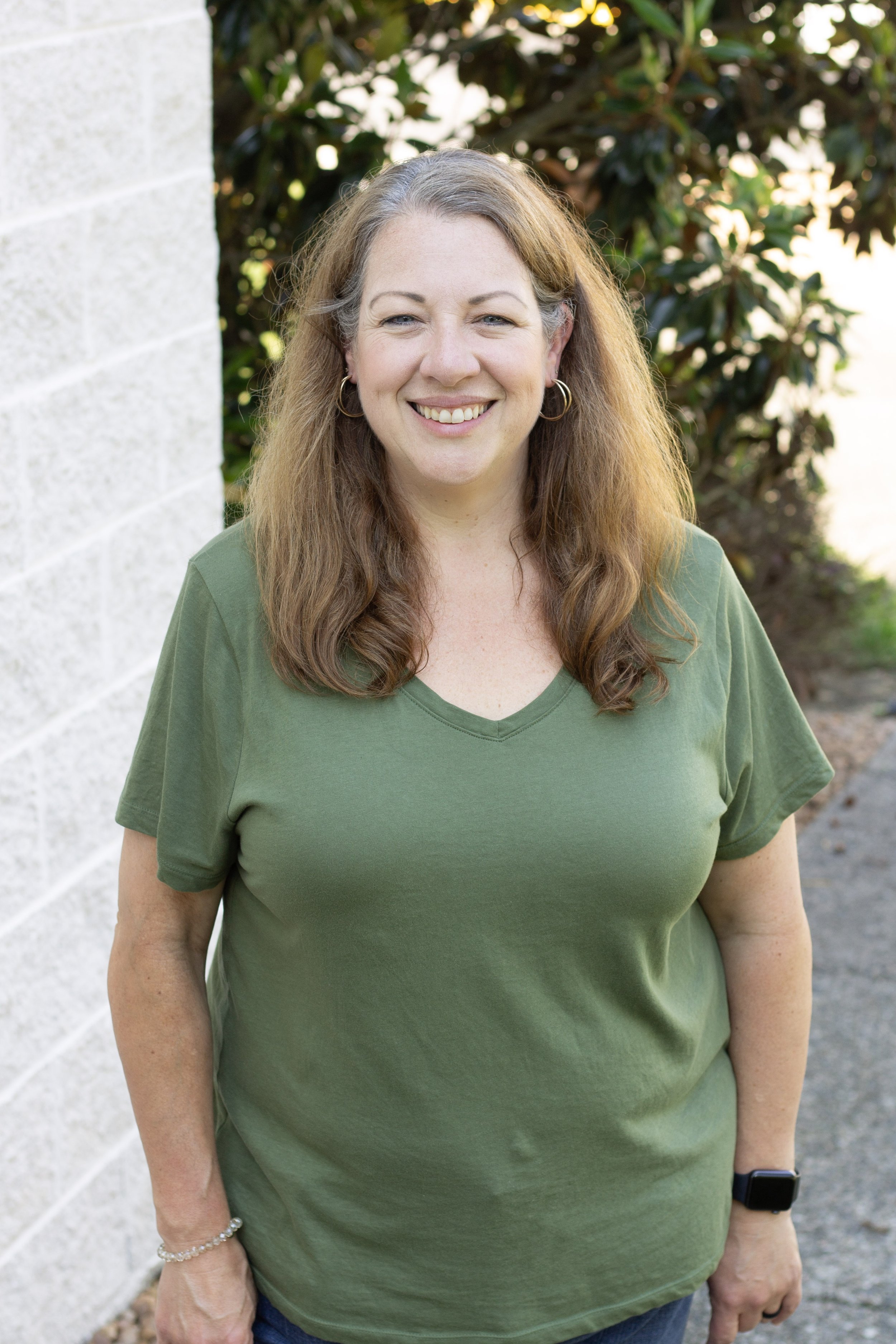 Smiling woman with long wavy hair wearing a green t-shirt, standing outdoors next to a white brick wall and green bushes.