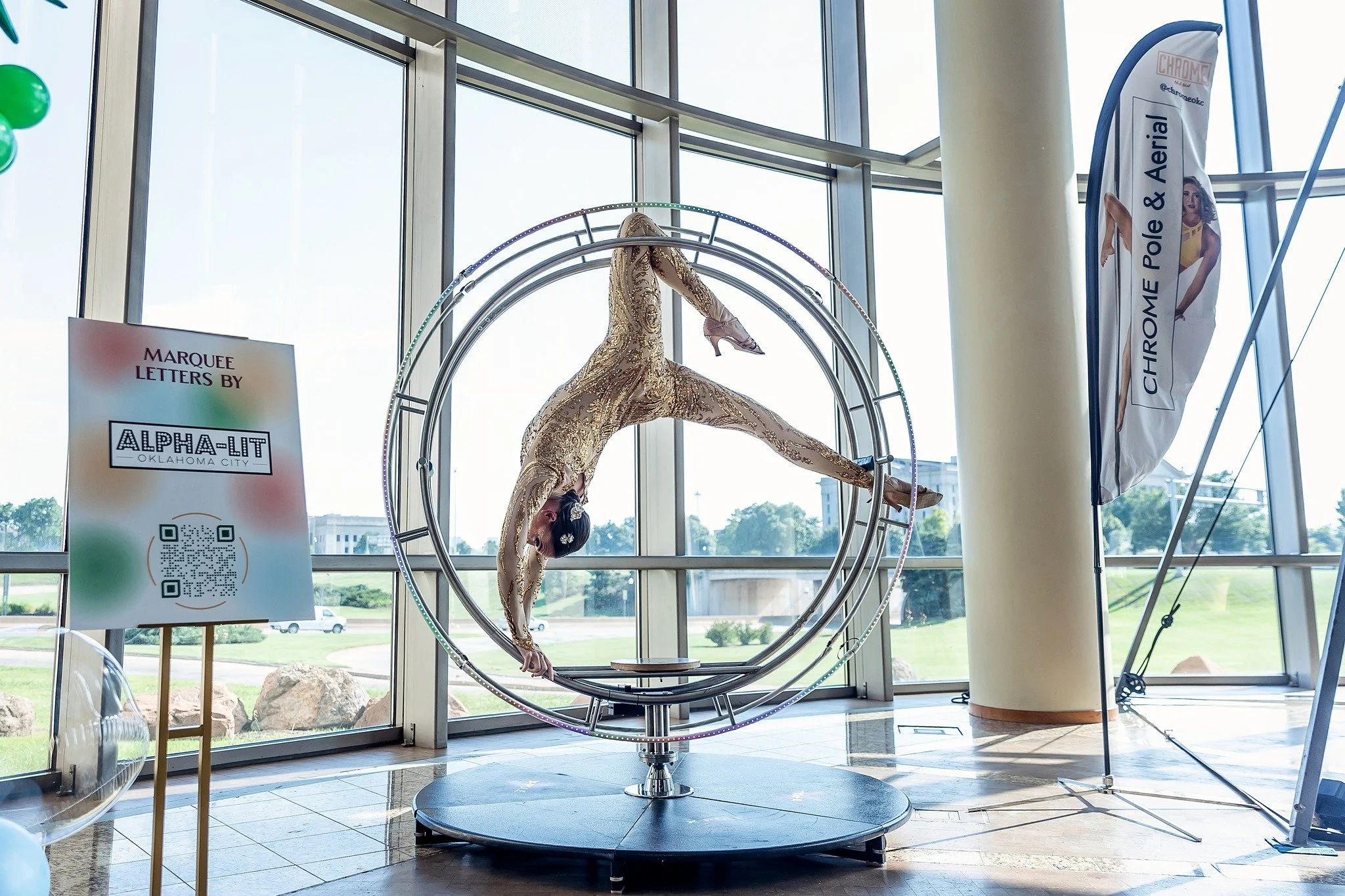 A gymnast wearing a gold, sparkly leotard performs an acrobatic pose on a circular apparatus with a large window and a banner advertising a pole and aerial show in the background.