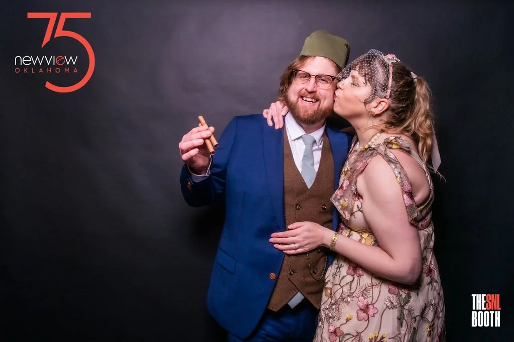 A man wearing a green hat and glasses, dressed in a blue jacket, with a woman in a floral dress and headband, kissing his cheek at a photo booth during an event celebrating 75 years of Newview Oklahoma.