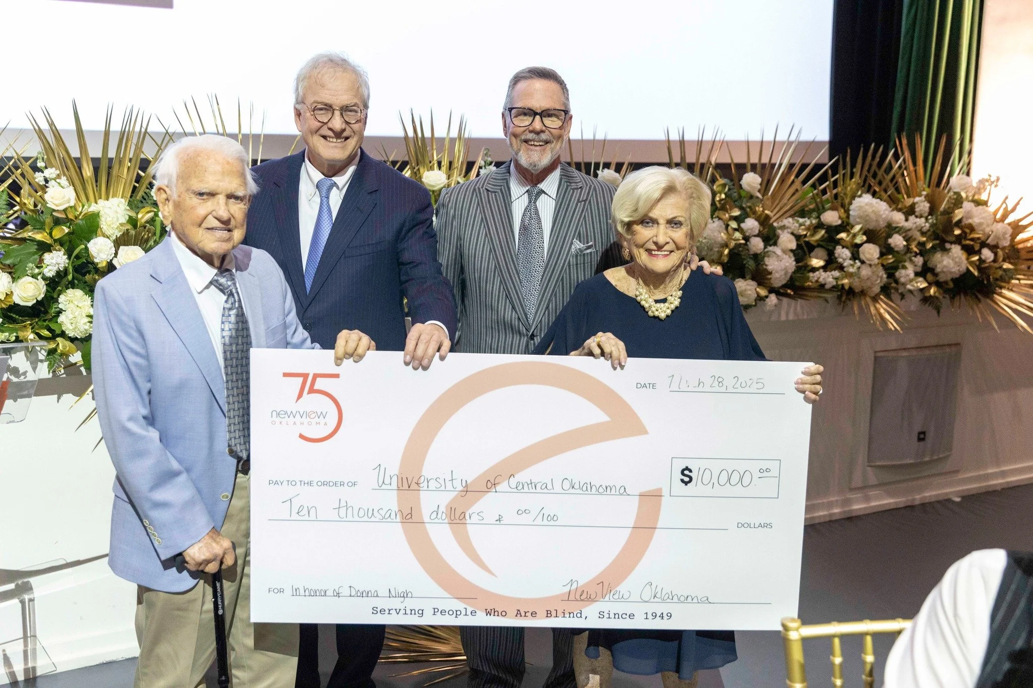 Five elderly individuals standing behind a ceremonial check for ten thousand dollars, with floral arrangements and a large white screen in the background. They are dressed formally, smiling, and appear to be at a formal event or presentation.