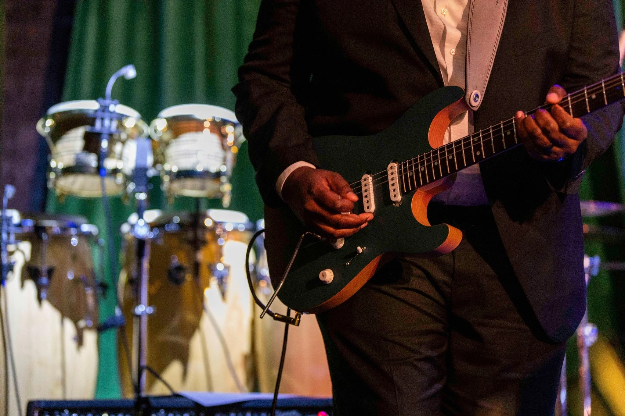 A person dressed in a suit playing an electric guitar, with percussion instruments visible in the background, possibly at a live performance or recording studio.