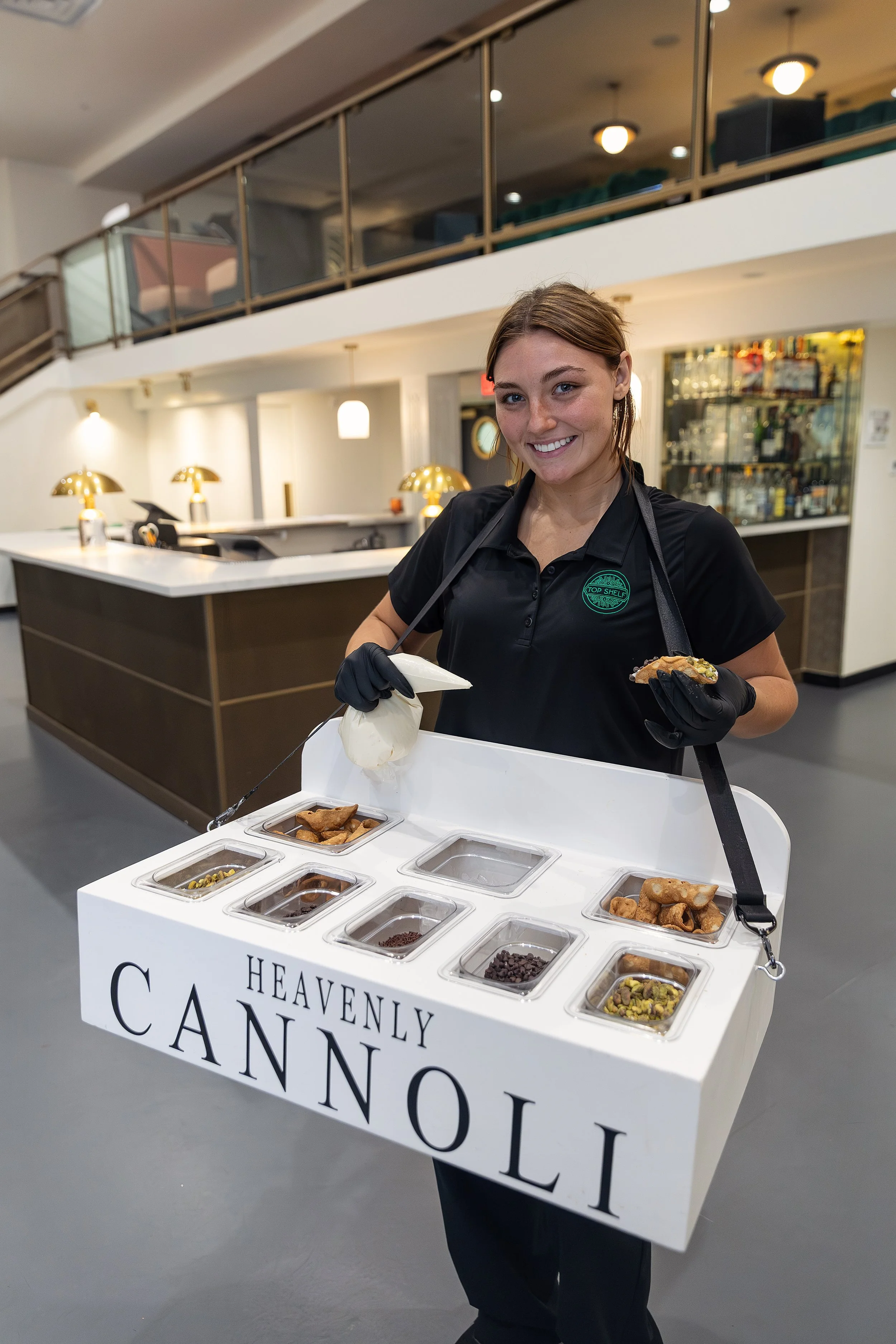 A woman wearing a black shirt and black gloves holding a white tray with multiple sections filled with different candies in a cafe or restaurant setting.